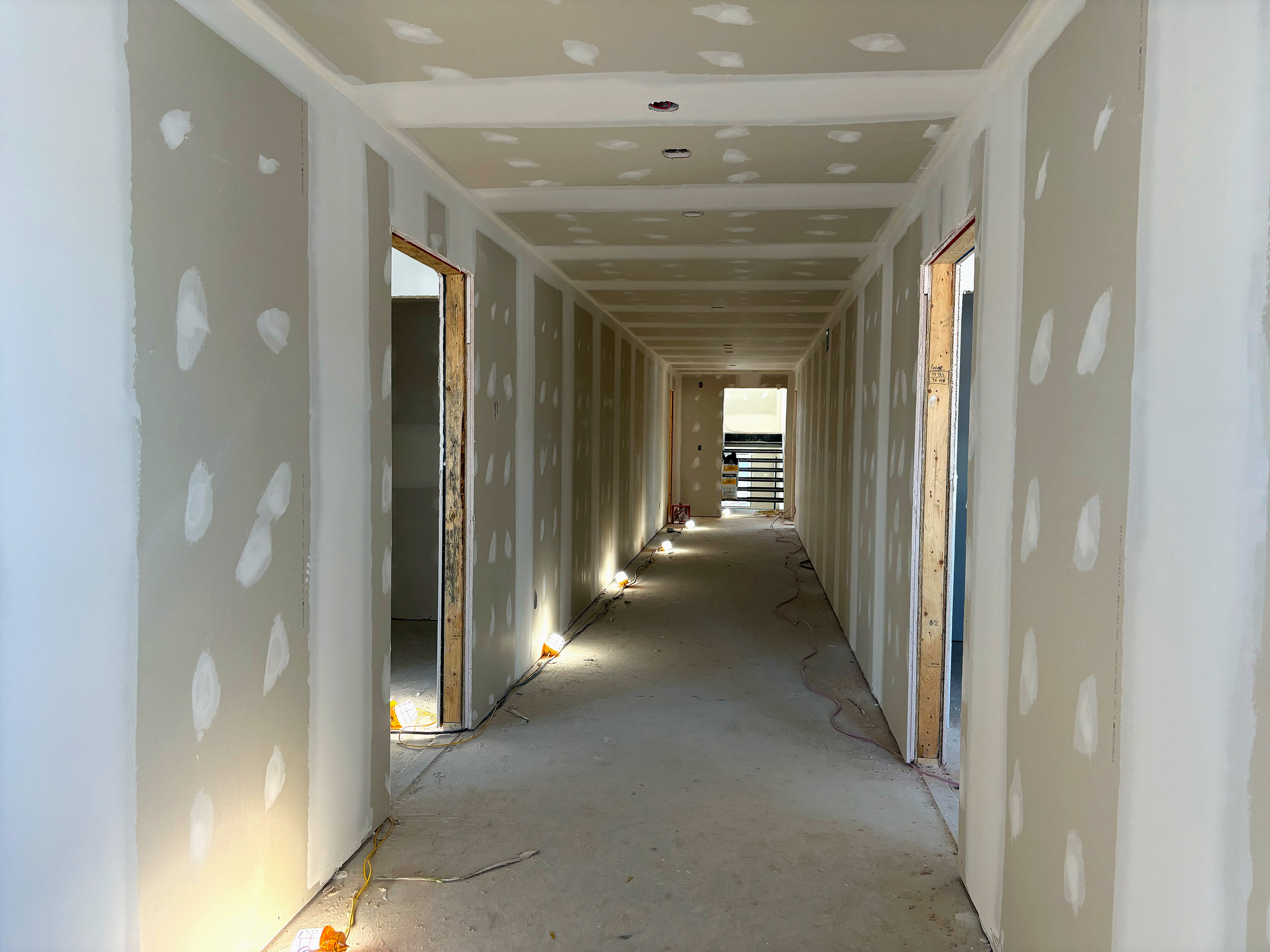 Couloir d'un bâtiment en construction avec murs et plafond en plaques de plâtre non peintes, éclairé par des lampes électriques posées au sol, avec plusieurs portes ouvertes le long du couloir et un balcon visible au fond.