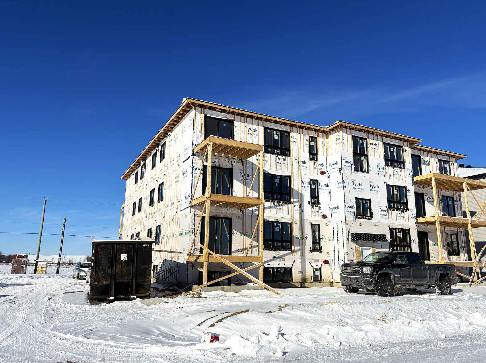 Construction d'un bâtiment résidentiel en hiver avec un ciel bleu, un camion noir, des poteaux électriques, et du neige au sol.