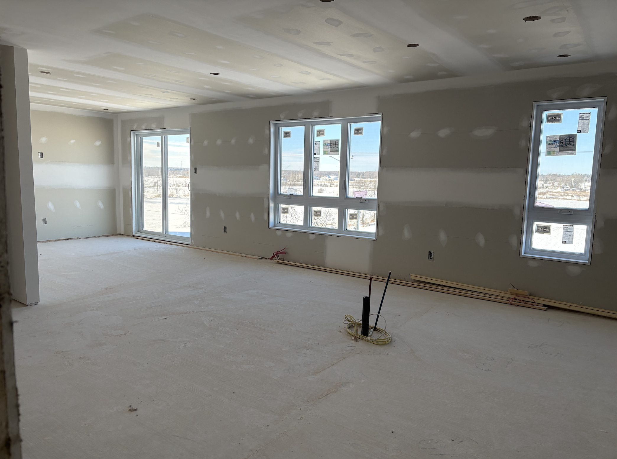 Une pièce de construction intérieure inachevée avec des murs recouverts de plaques de plâtre, plusieurs fenêtres laissant passer la lumière naturelle, un sol en béton brut, et des câbles électriques sortant du sol.