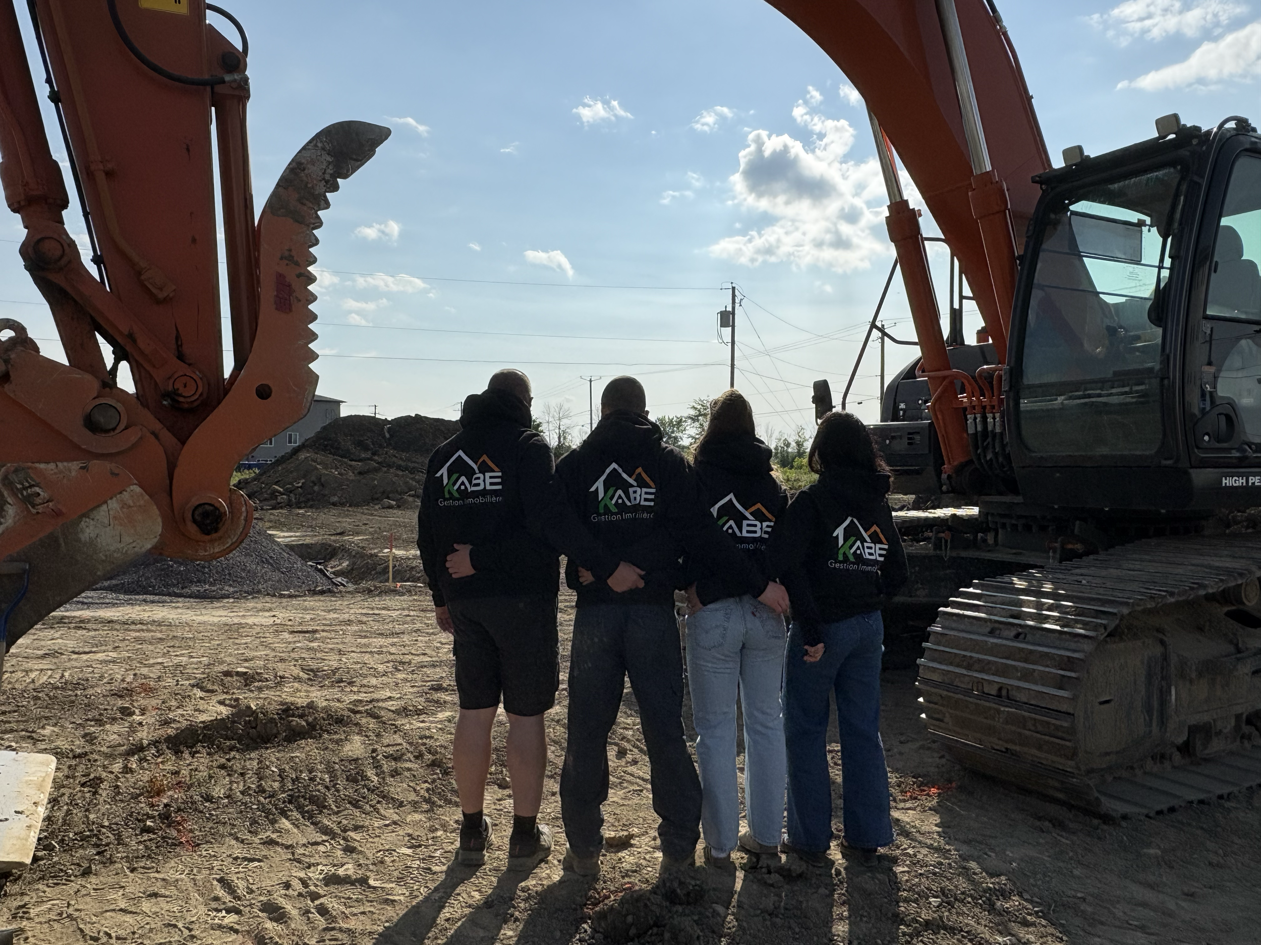 quatre personnes portant des sweats à capuche avec un logo KABE debout à côté d'une excavatrice sur un site de construction.