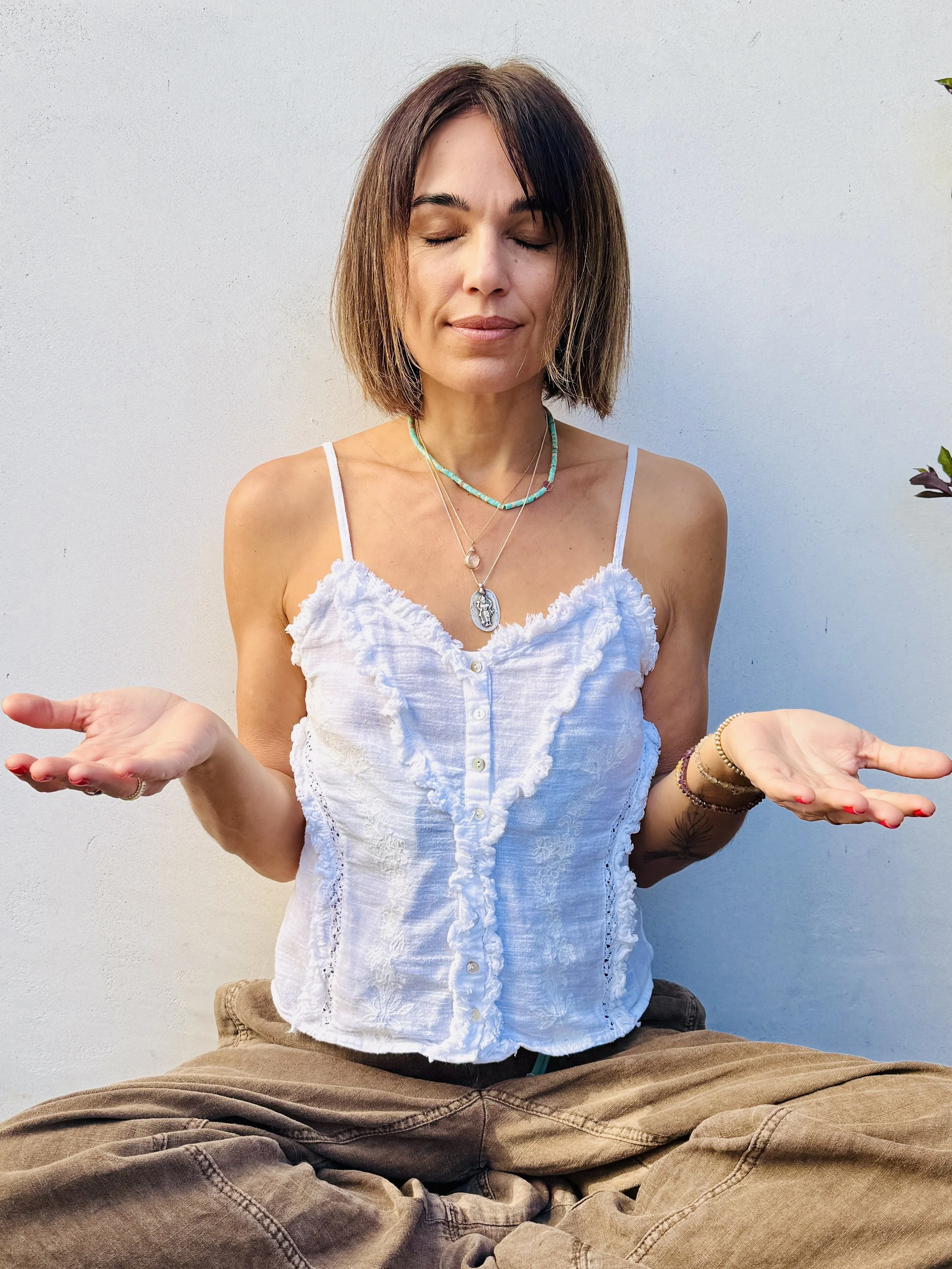 Indra Kelly with shoulder-length brown hair and closed eyes, sitting cross-legged against a plain light-colored wall, wearing a white sleeveless top and jewelry, with her palms facing upward.