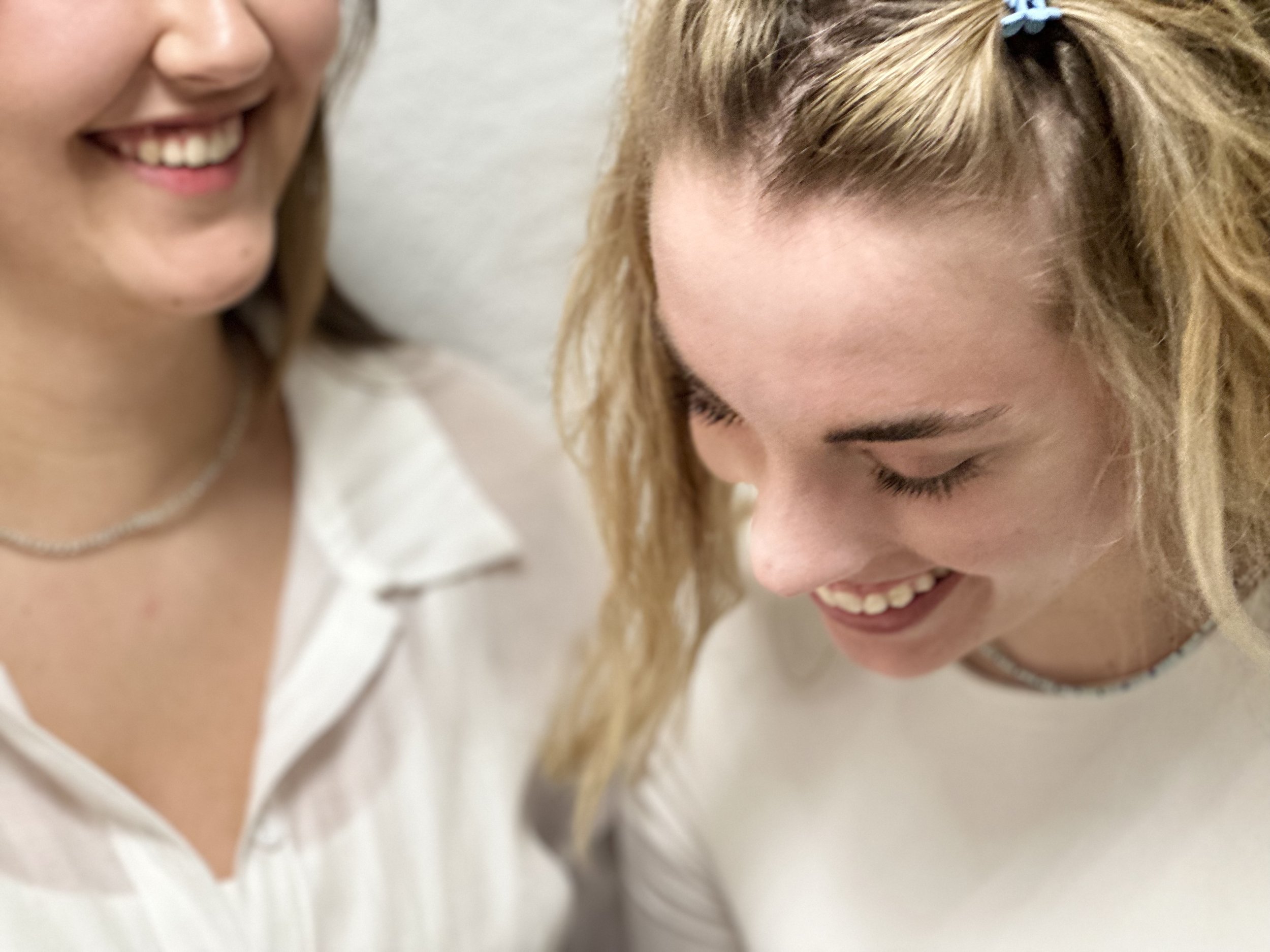 Zwei lachende Frauen, eine mit blondem, lockigem Haar, und eine mit braunem Haar, in weißen Oberteilen, beim Lachen.
