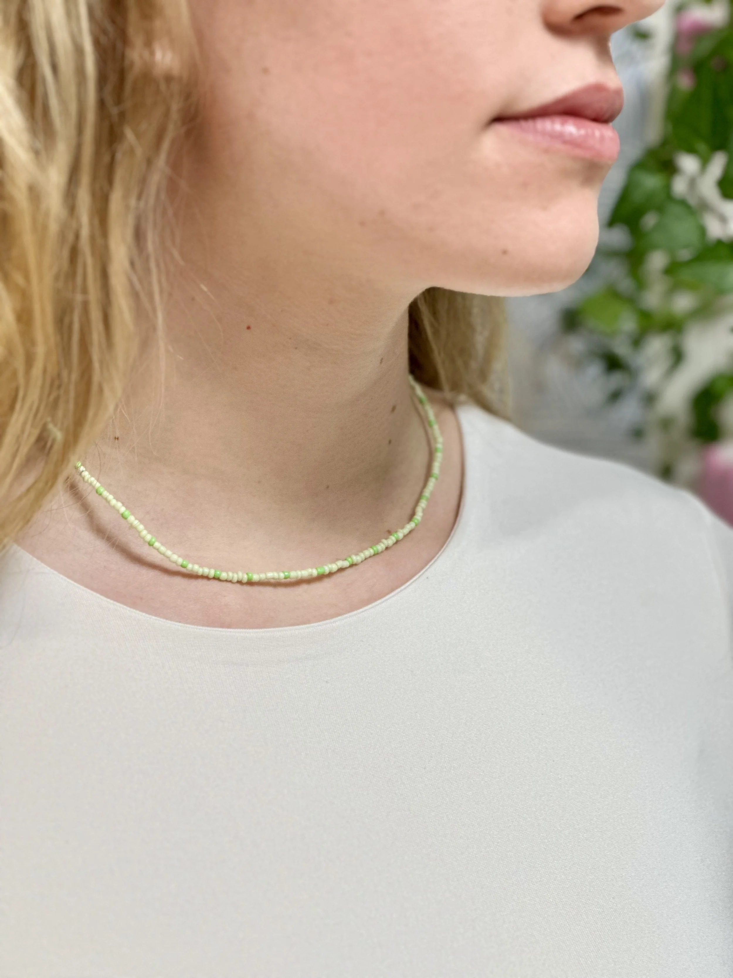 Nahaufnahme eines Mädchens mit heller Haut, blonden, lockigen Haaren, das eine dezente grüne Perlenkette trägt, vor einer blühenden Pflanze im Hintergrund.