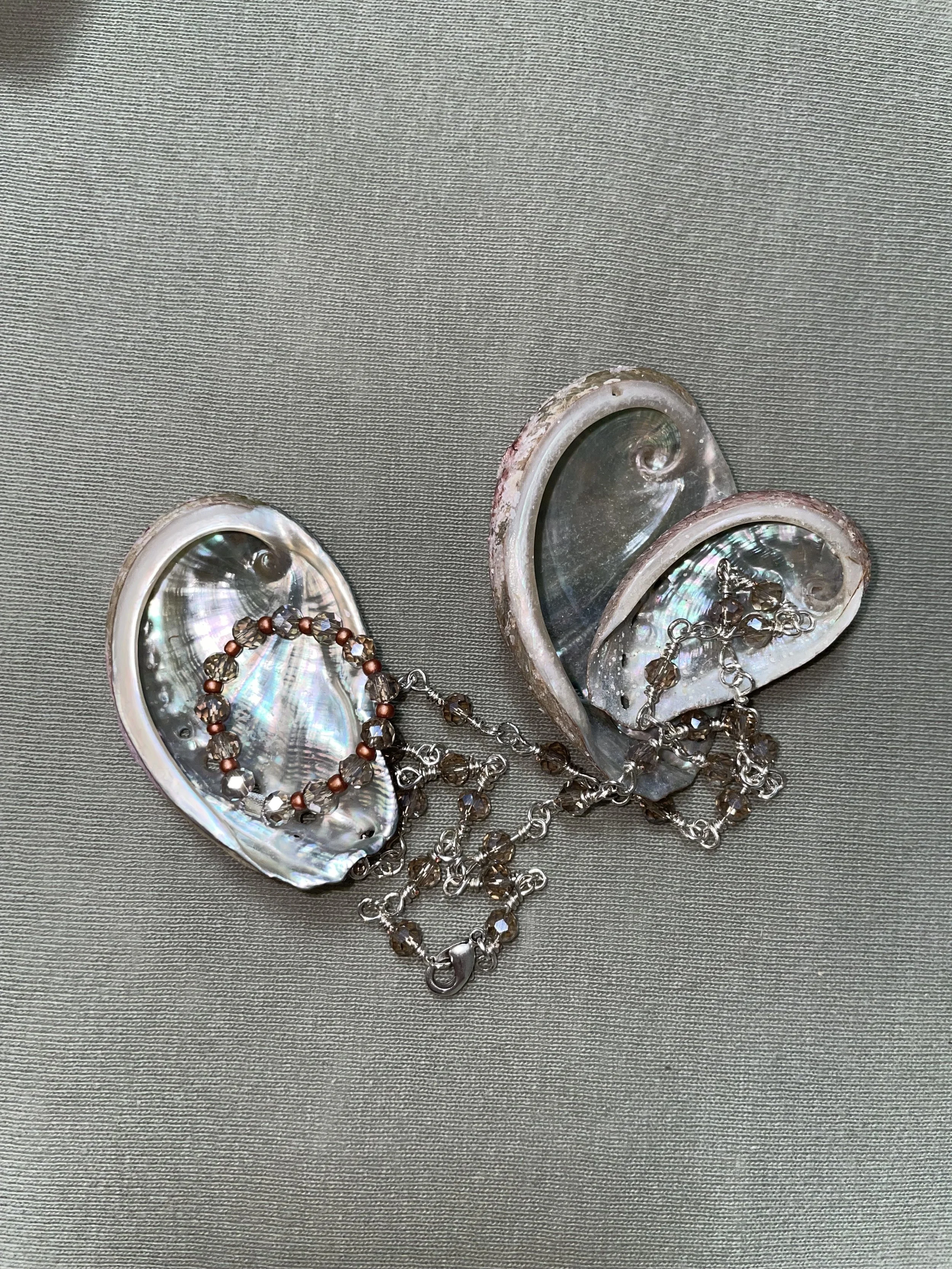 Zwei Muschelschalen, die zu Herzformen angeordnet sind, verbunden durch eine Kette mit Schmuckstücken, auf einem grauen Stoffhintergrund.