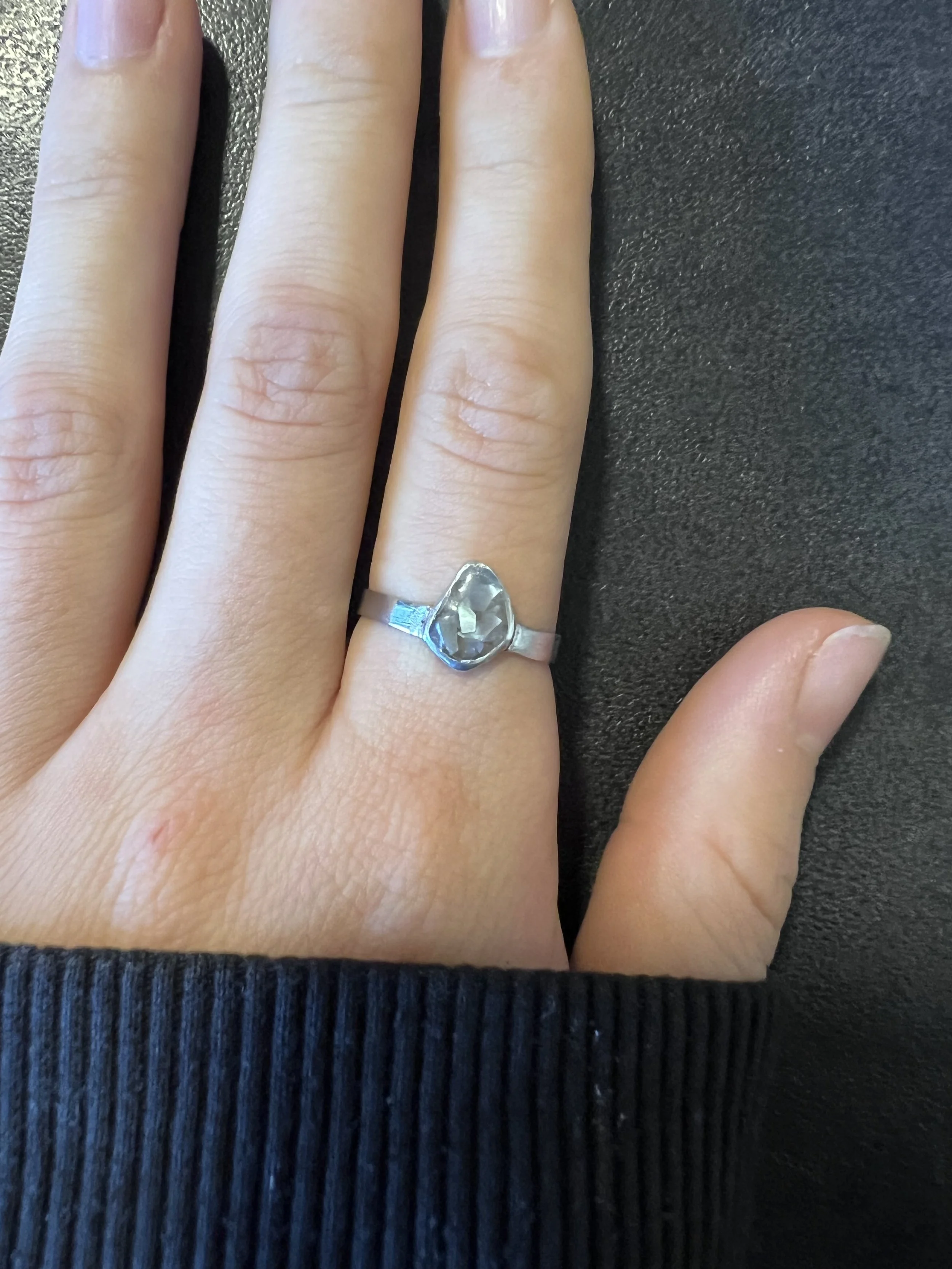 Eine Hand mit einem Silberring, der einen großen, transparenten, gefassten Edelstein trägt, liegt auf einer schwarzen Oberfläche.