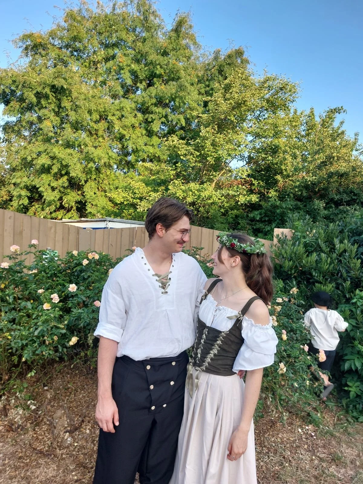 Ein Paar in traditioneller Kleidung schaut sich liebevoll an, im Hintergrund ein Garten mit grünen Büschen und Blumen, ein Zaun und ein bisschen Himmel.