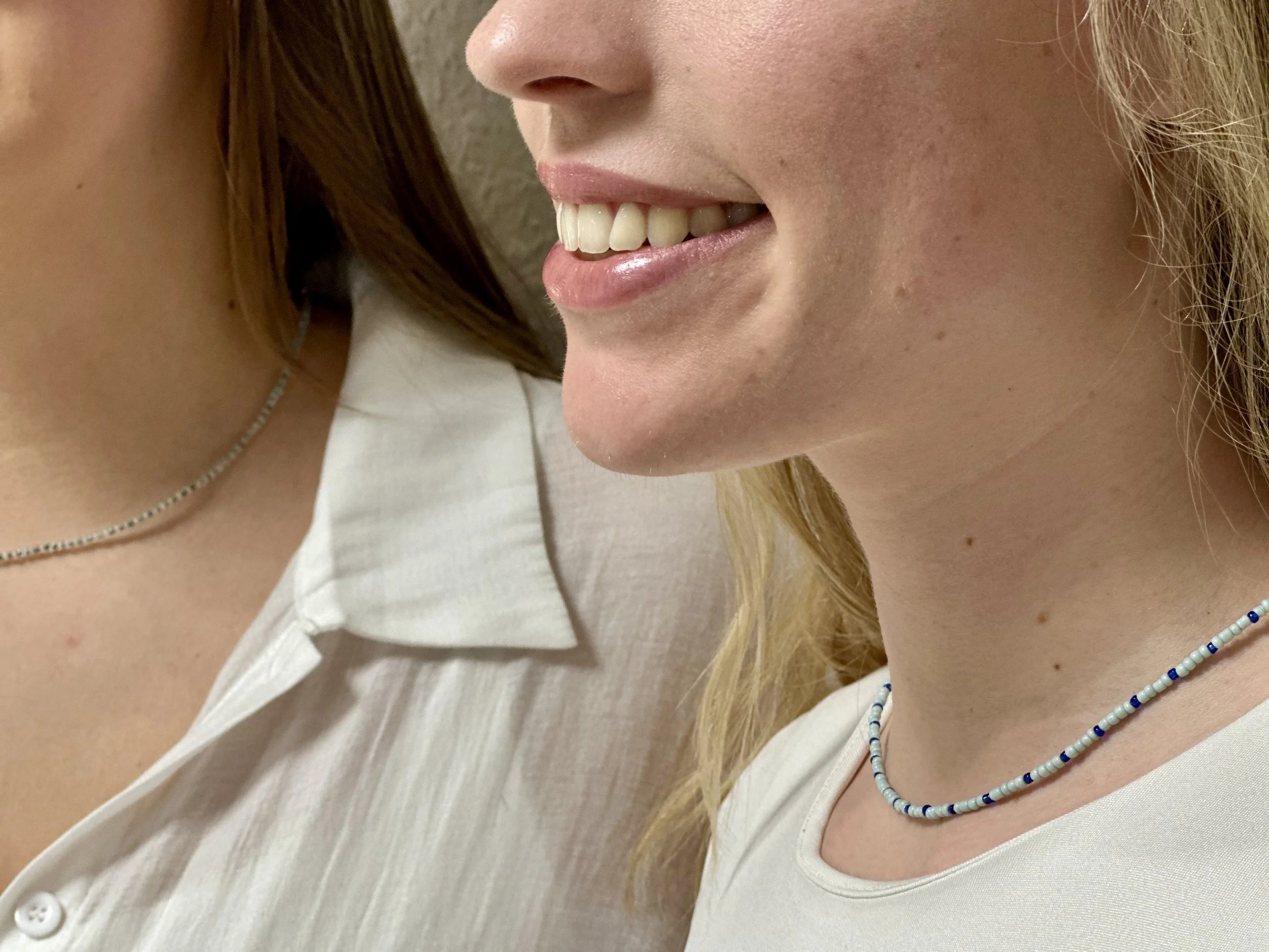 Nahaufnahme von zwei Menschen mit weißen Oberteilen, einer trägt eine blaue Perlenkette, lächelt und zeigt nur einen Teil ihres Gesichts und Halses.