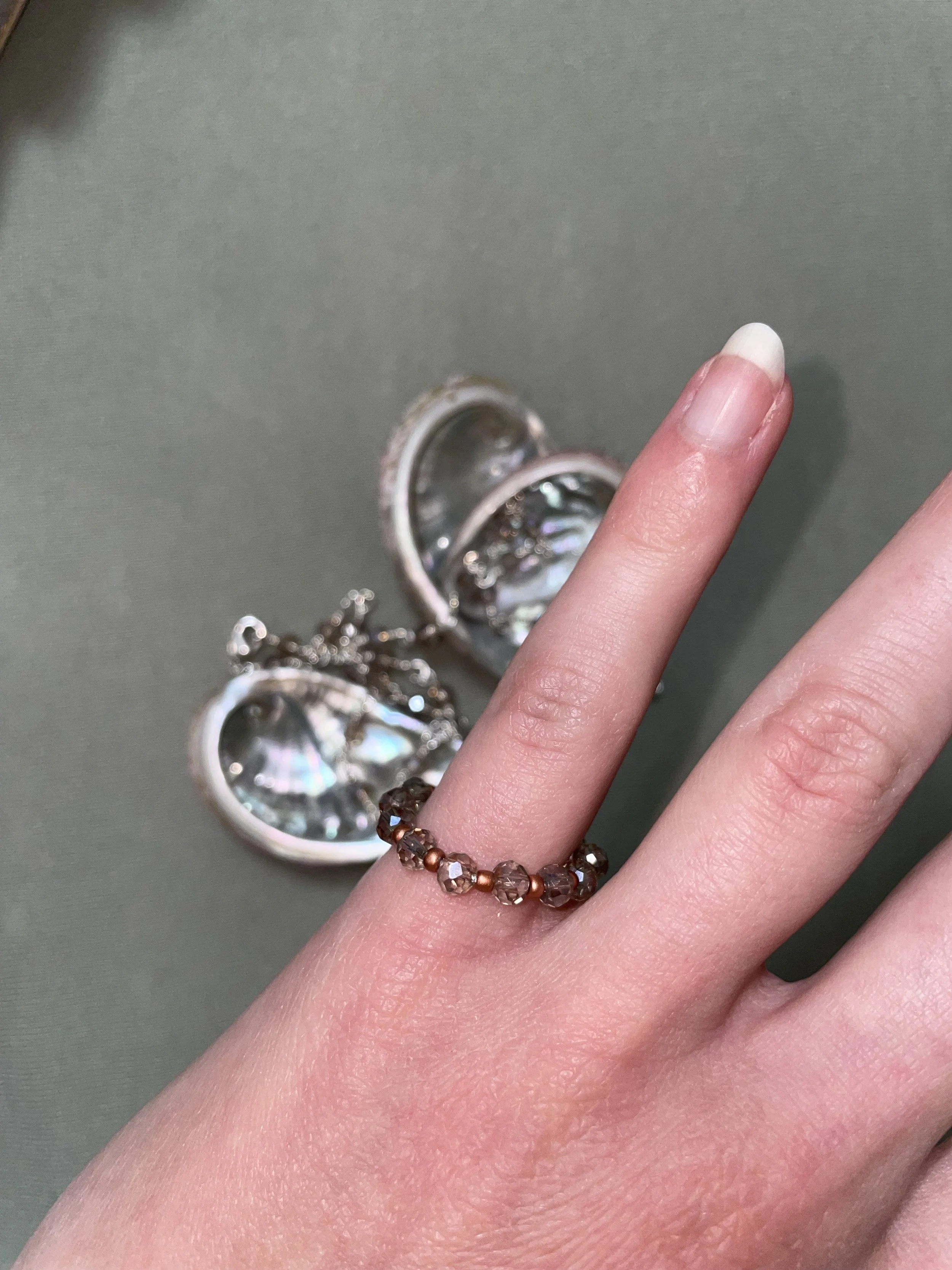 Die Hand trägt einen braunen Perlenring, im Hintergrund liegen offene Muschelschmuckstücke auf einem grauen Untergrund.