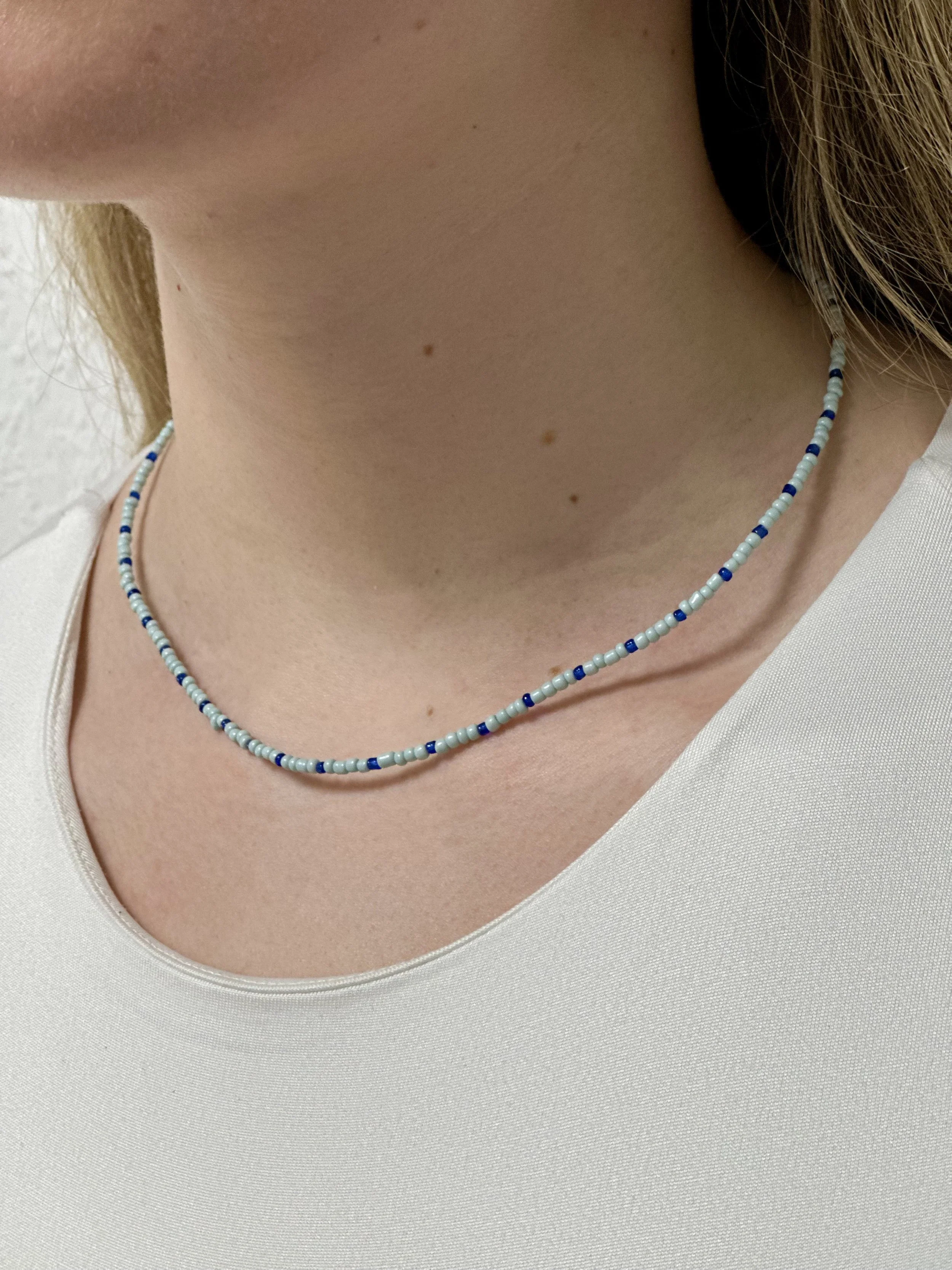 Nahaufnahme eines Halses mit einer Perlenkette, die hauptsächlich aus weißen und blauen Perlen besteht, getragen von einer Person in einem weißen Oberteil.