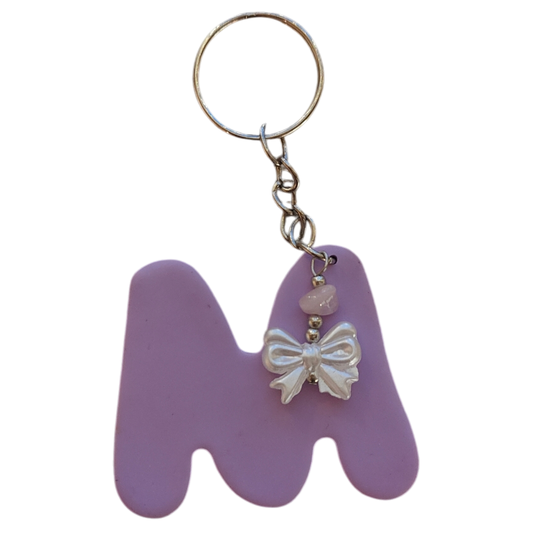 Lila avaimenperä, jossa iso kirjaimen M muotoinen koriste ja pieni valkoinen rusetti. Avaimenperä omalla nimikirjaimella itsetehty korupaja sodankylä lappi kaadodesign