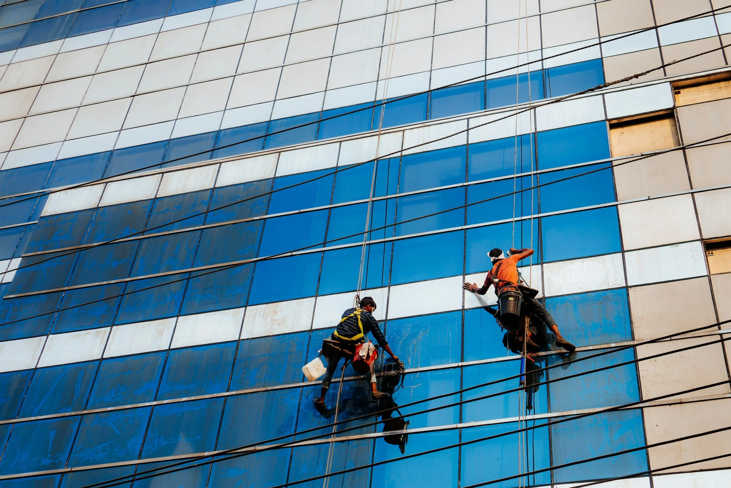 Two window cleaners are suspended on ropes cleaning the glass windows of a tall building with a modern glass facade.