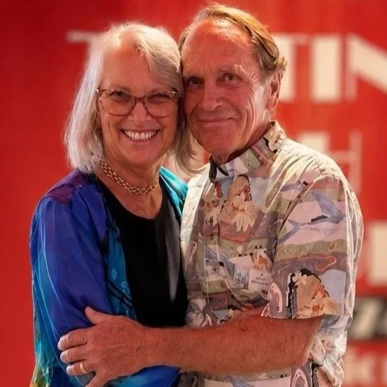A smiling older woman with white hair and glasses, wearing a blue jacket, hugging a smiling man with light brown hair in a patterned shirt, against a red background.