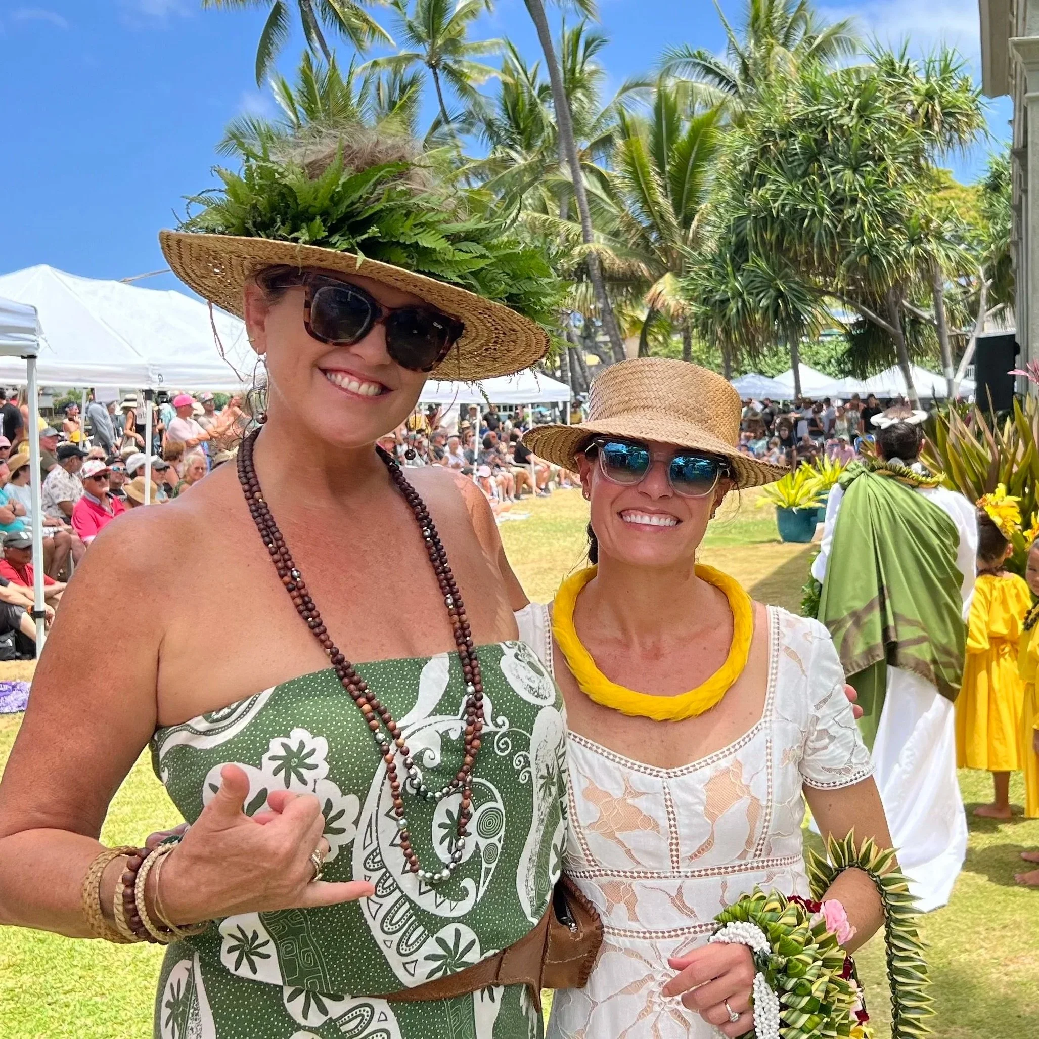 Two women at an outdoor event wearing straw hats and sunglasses, with a crowd of people and palm trees in the background.