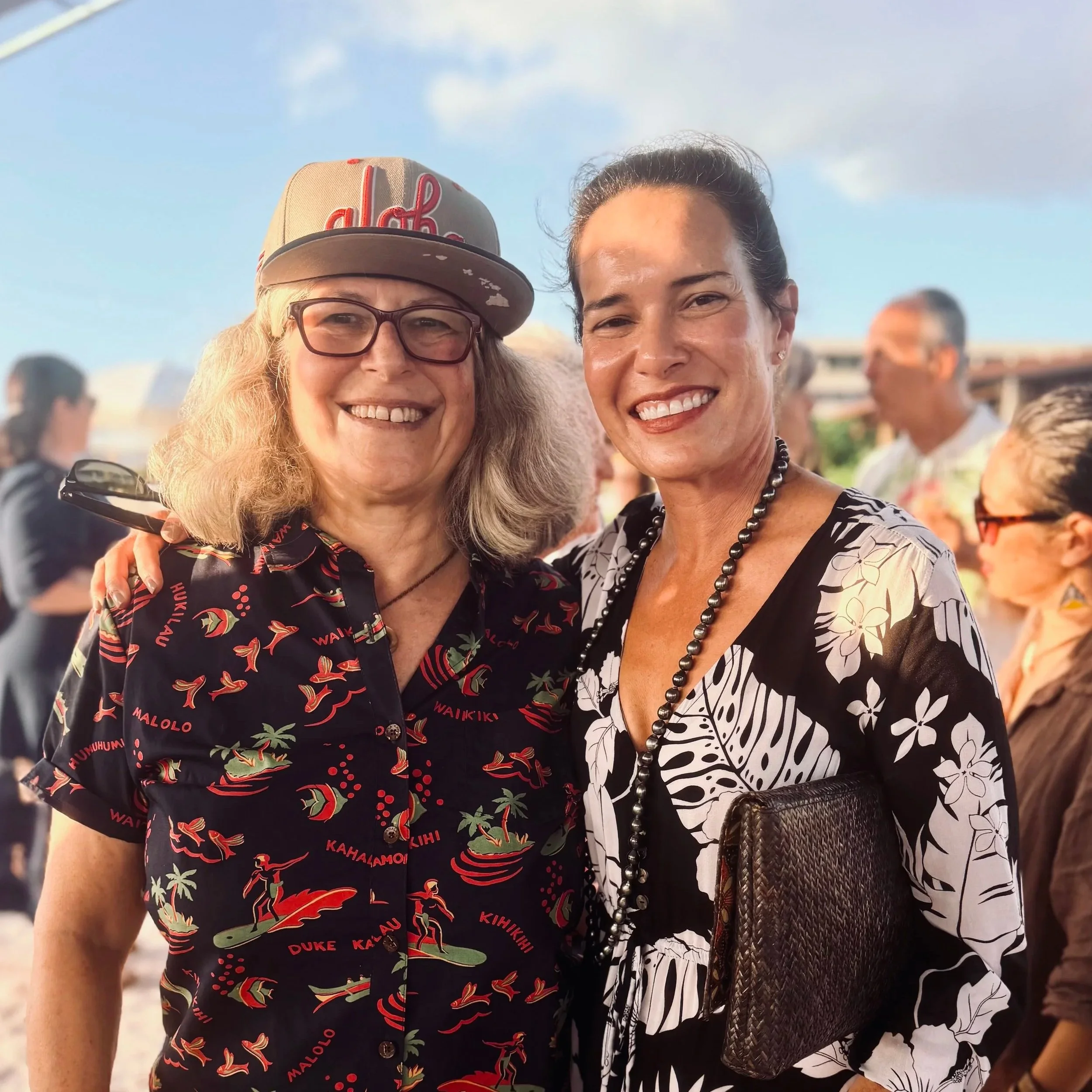 Two women smiling at an outdoor event, one wearing a beige cap with red embroidery and glasses, the other with dark hair pulled back, wearing a black and white floral dress, necklace, and holding a brown handbag.