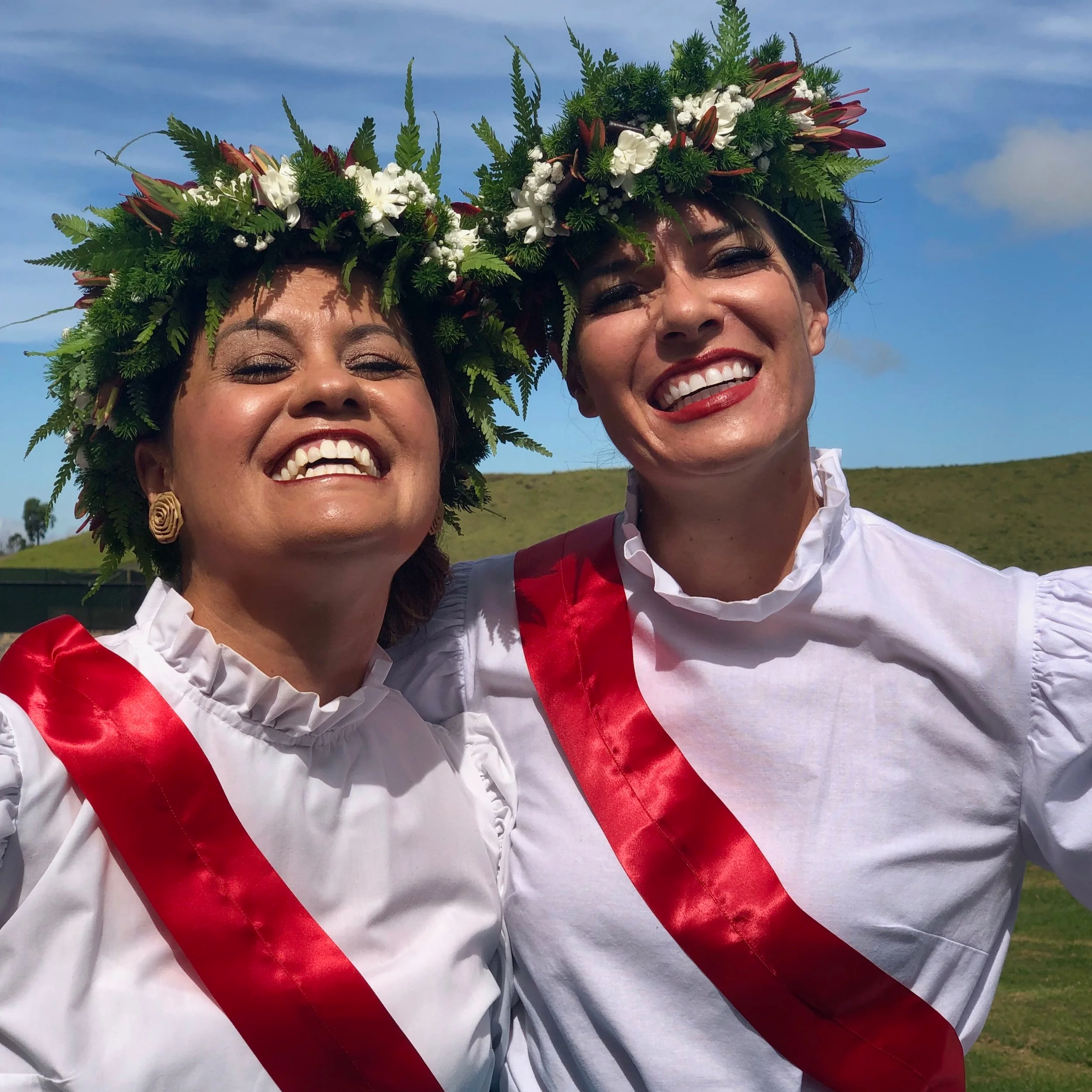 Two women smiling, wearing white dresses with red sashes and floral crowns made of green ferns, white flowers, and burgundy accents, outdoors against a blue sky.