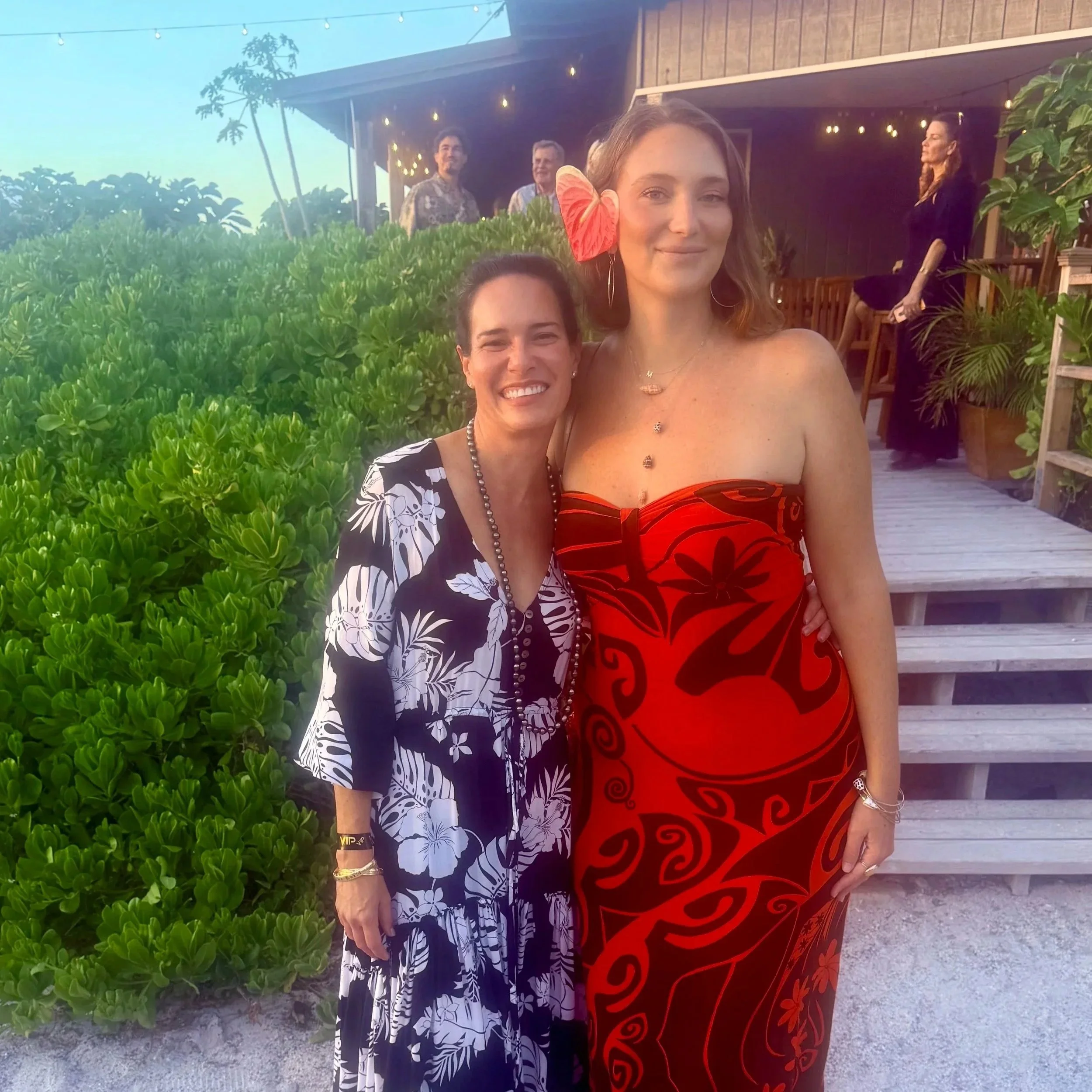 Two women standing outdoors at sunset, one with a red flower in her hair and wearing a red dress, the other in a black and white floral dress, with three men in the background near a wooden structure decorated with string lights.