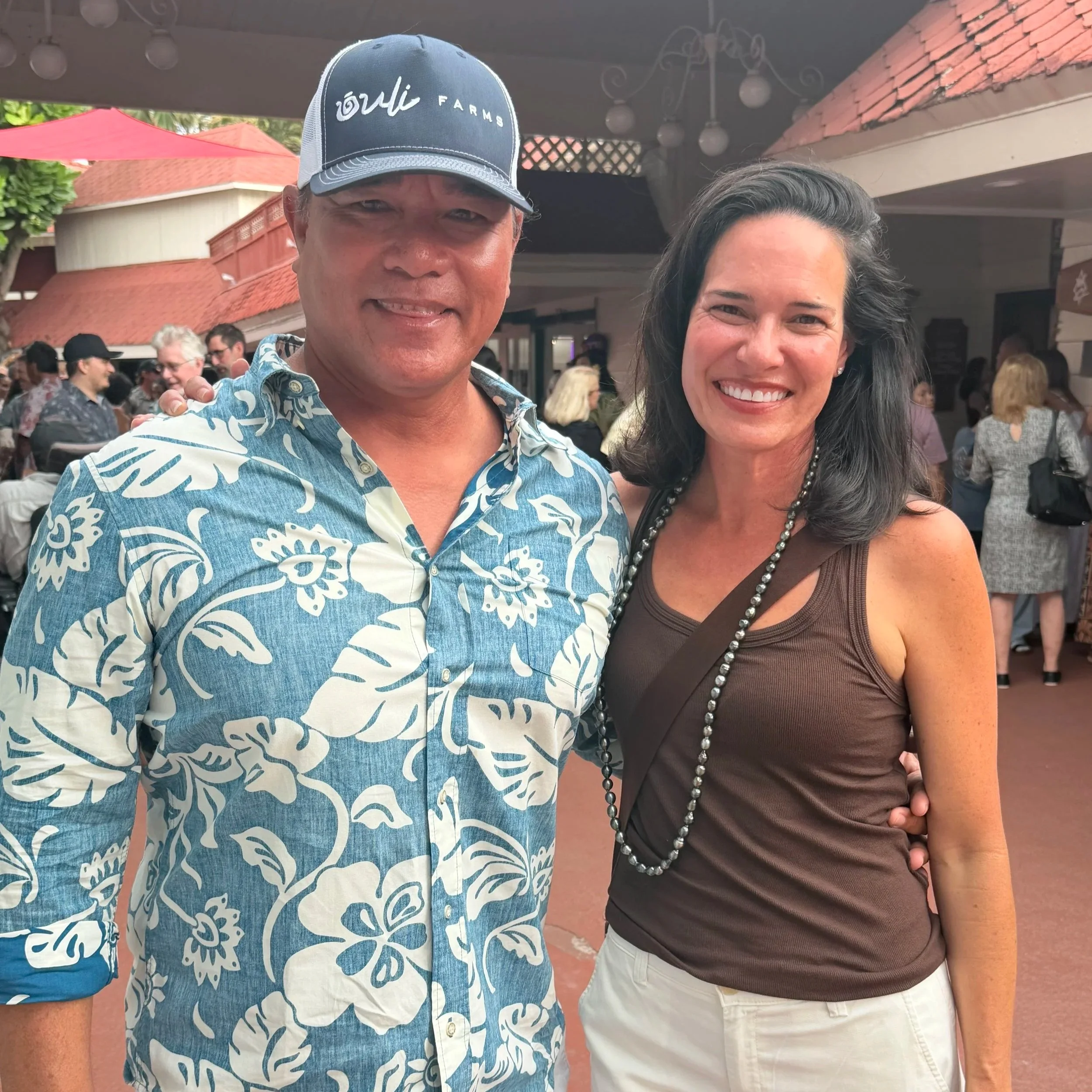 A man in a blue Hawaiian shirt and a baseball cap standing next to a woman in a brown shirt and white pants at an outdoor venue with a crowd in the background.