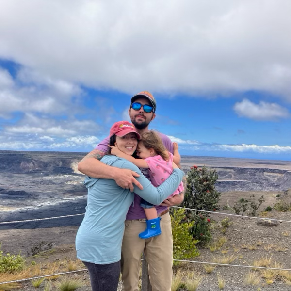 A family of three hugging outdoors in a scenic volcanic landscape with a cloudy sky.