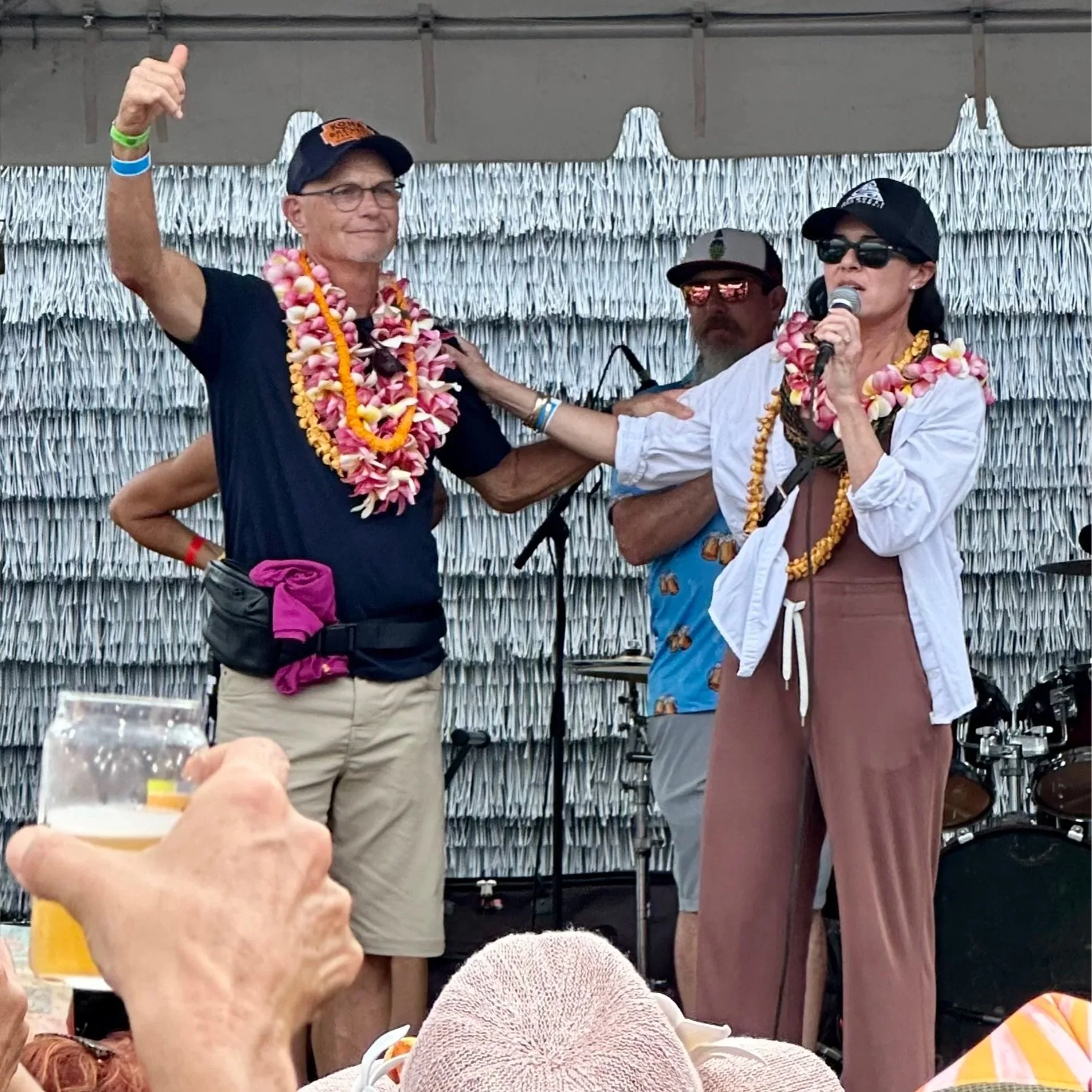 People celebrating on a stage decorated with white fringe, with floral leis and colorful clothing, and one person speaking into a microphone.