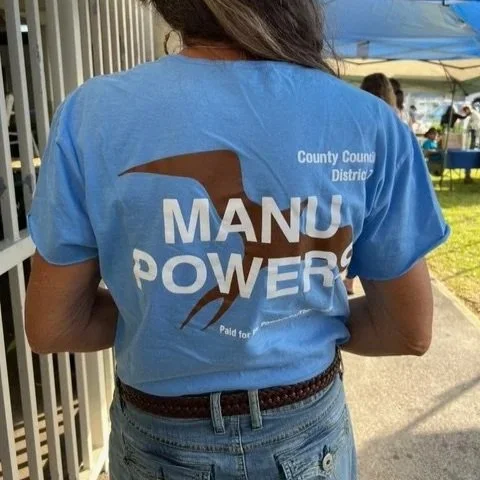 A woman wearing a blue T-shirt with the text "MANU POWER" and a stylized bird logo on the back, standing outdoors near a fence and under a tent.