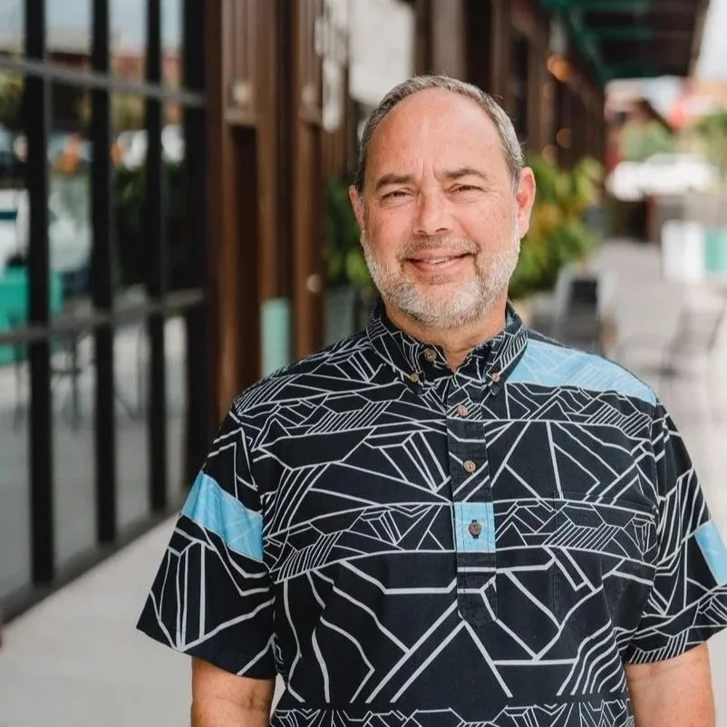 A middle-aged man with gray hair and a beard stands outside a building with large windows, smiling at the camera, wearing a black shirt with a geometric pattern and blue accents.