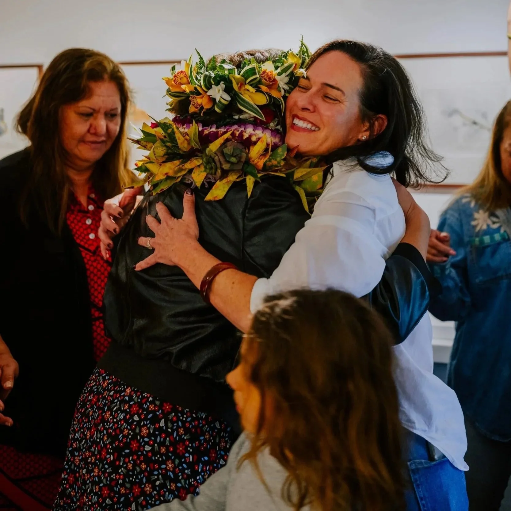 Two women hugging, one woman smiling, wearing a white shirt, and another woman with floral scarf, holding a bouquet of flowers. Other women and children are present in the background.