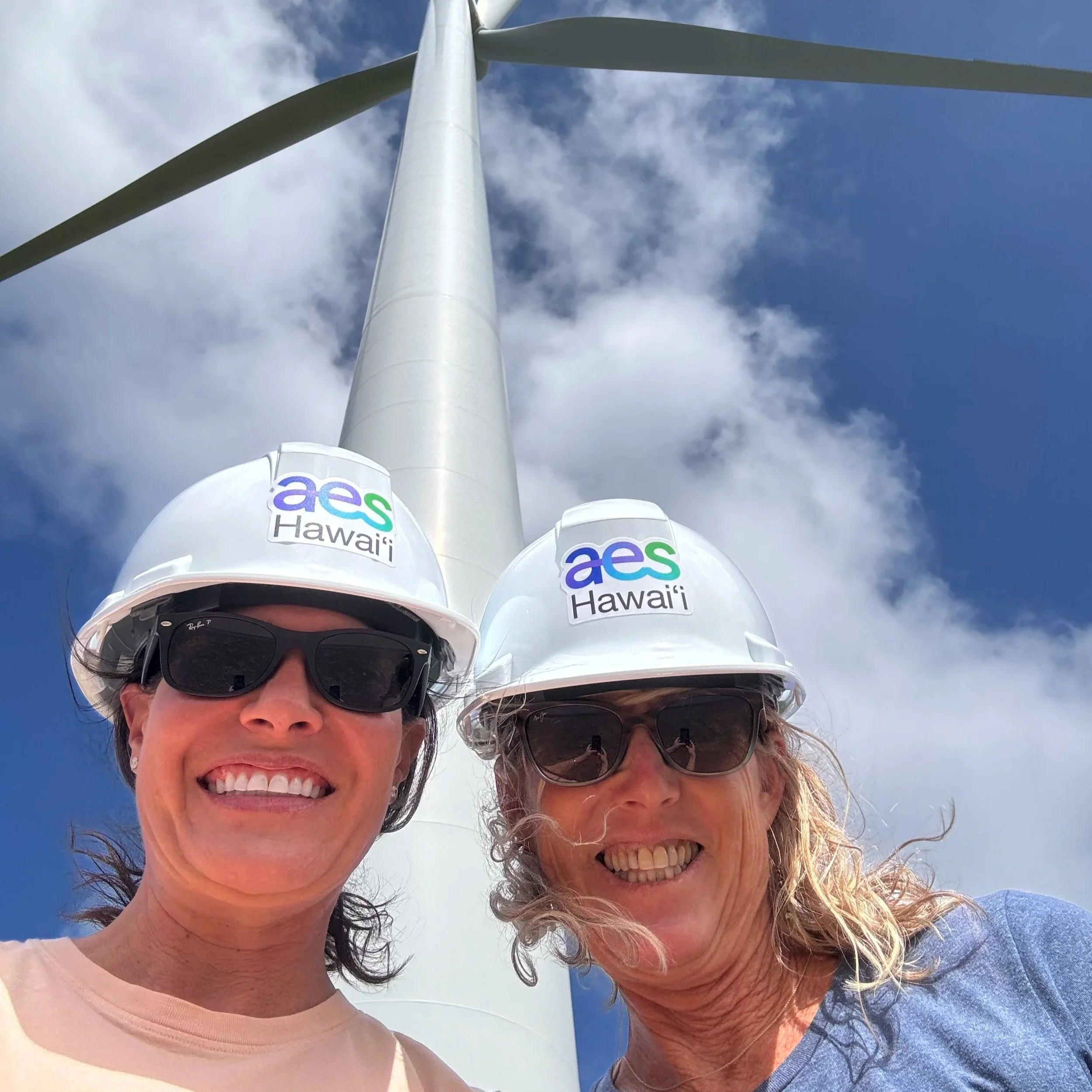Two women smiling, taking a selfie outdoors with wind turbines and a partly cloudy sky in the background.