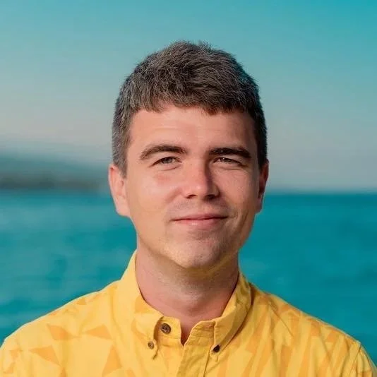A young man with short brown hair smiling, wearing a yellow shirt, standing outdoors with the ocean and sky in the background.