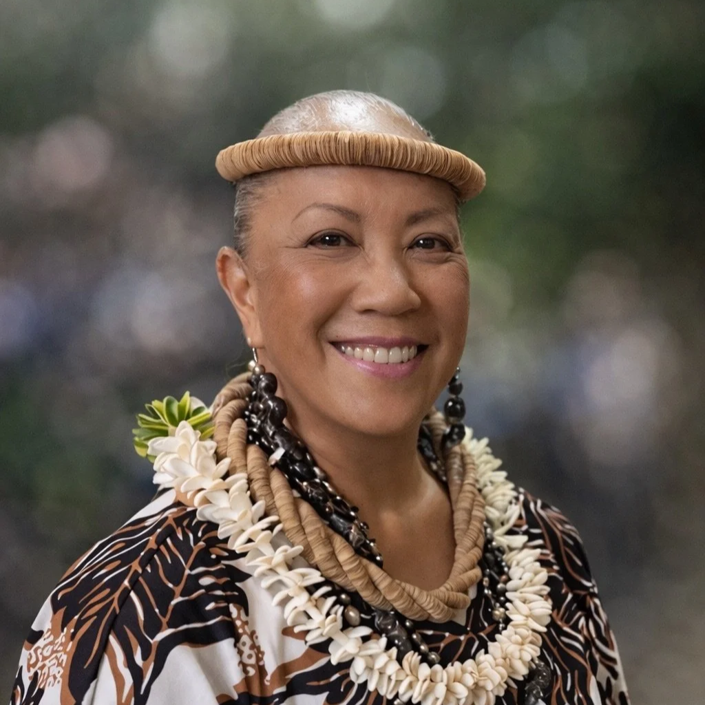 A smiling woman wearing traditional Polynesian jewelry, including a circular headband, multiple necklaces, and earrings, with a patterned garment against a blurred outdoor background.