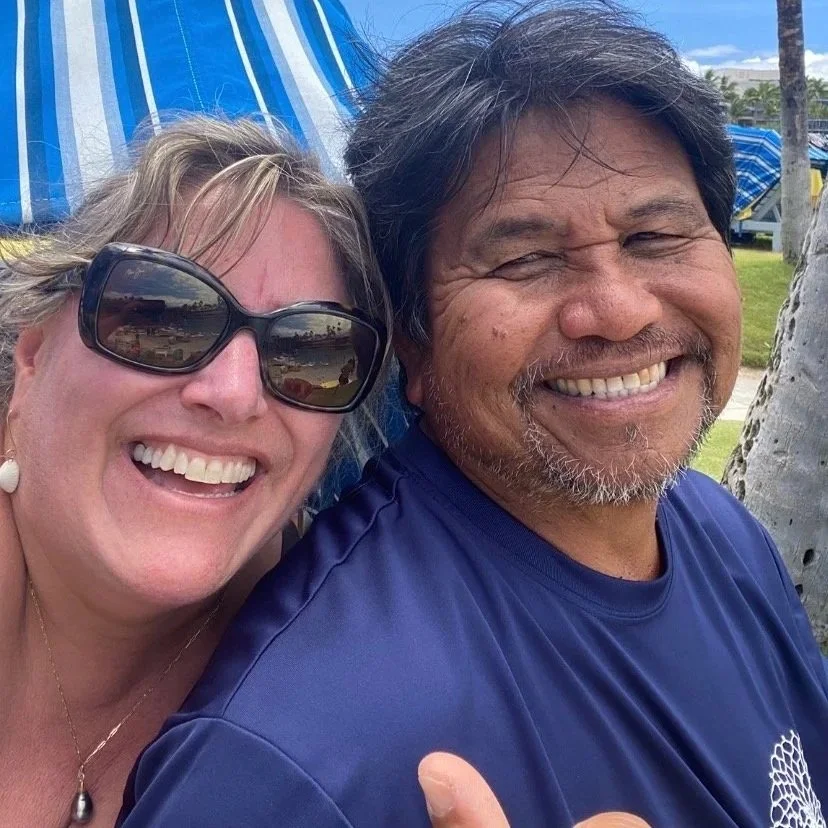 A smiling woman wearing sunglasses and earrings, and a smiling man with facial hair, are taking a selfie outdoors on a sunny day. There are blue and white striped structures and palm trees in the background.