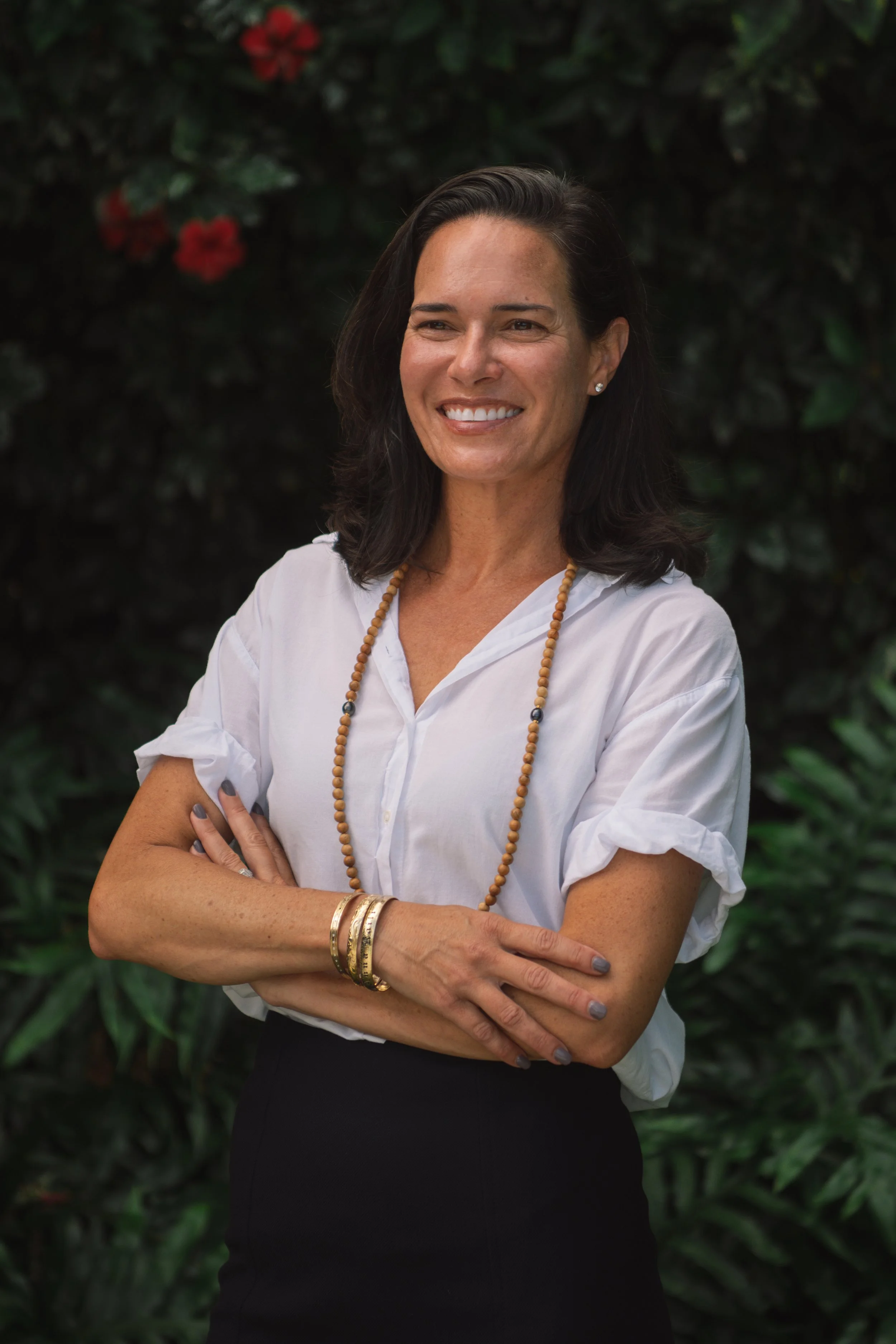 Hawaii County Council District 7 Candidate standing outdoors with her arms crossed, smiling, wearing a white blouse, black skirt, and jewelry including a long beaded necklace, bangles, and earrings, with greenery and red flowers in the background.