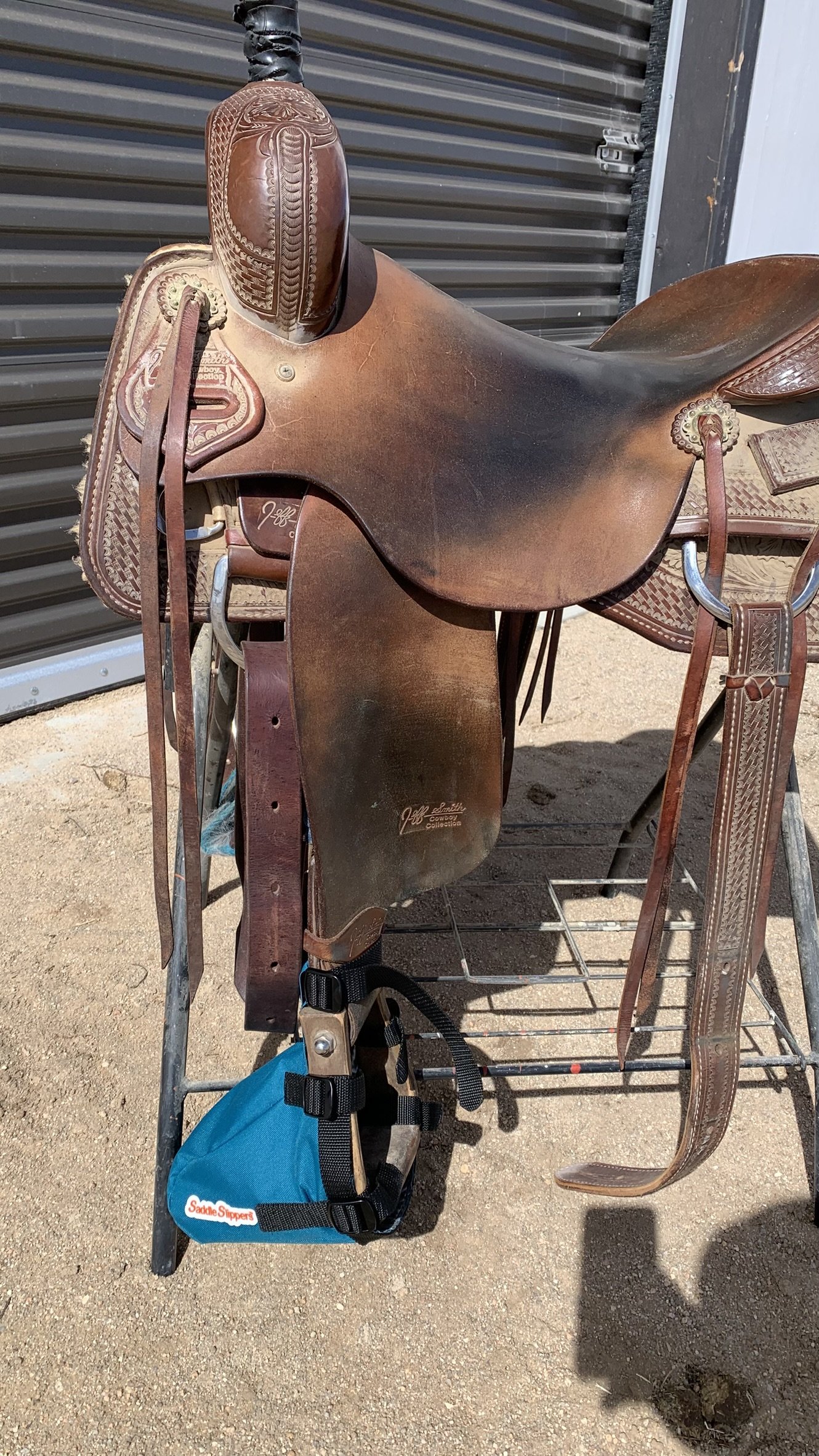 A Western-style brown leather saddle, resting on a metal saddle stand with a turquoise Saddle Slipper attached to the stirrup.