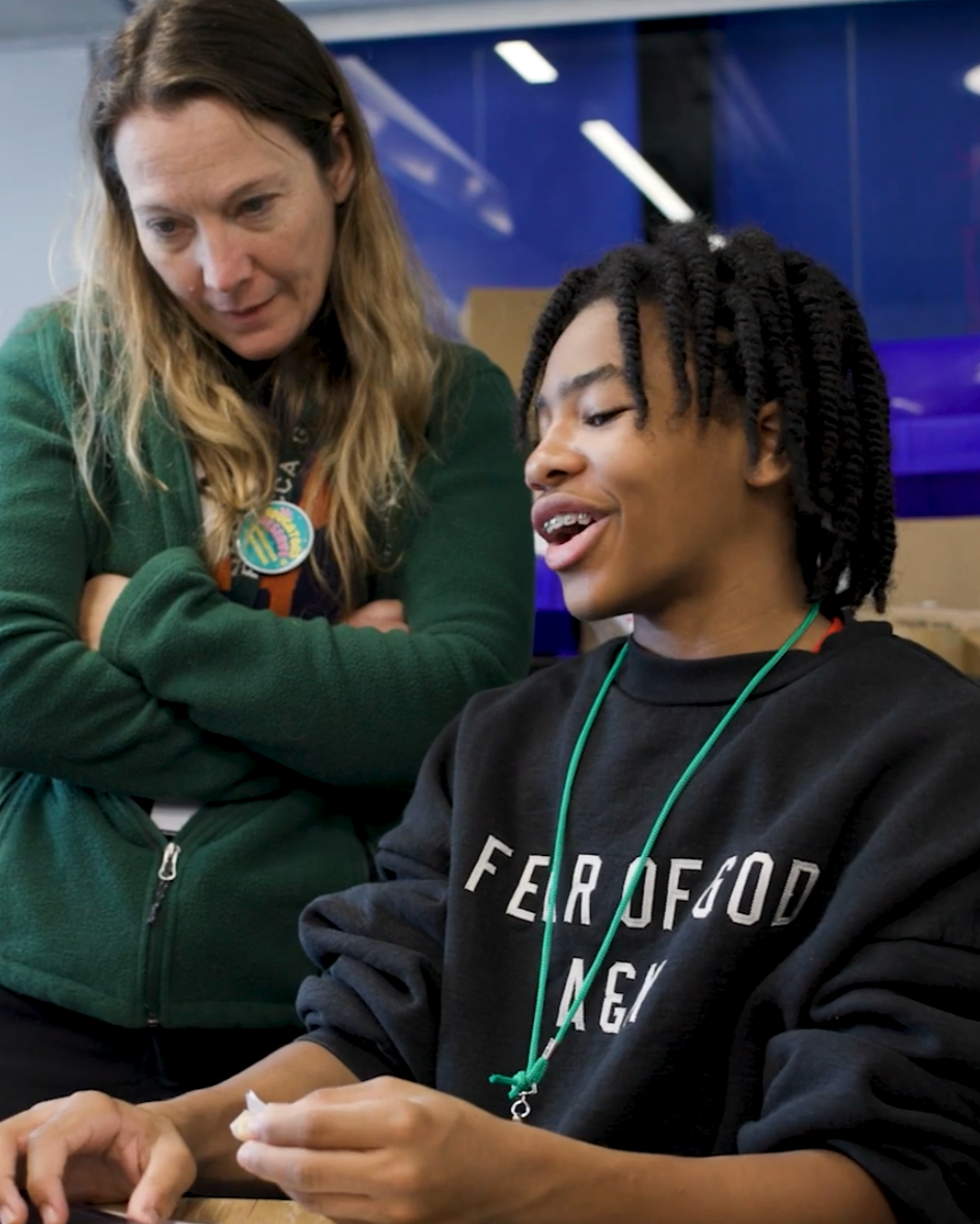 A woman with long, wavy blonde hair wearing a green jacket and a lanyard is looking at a young man with braided black hair who is wearing a black sweatshirt with white text and a green lanyard. They are indoors, engaging in a conversation.