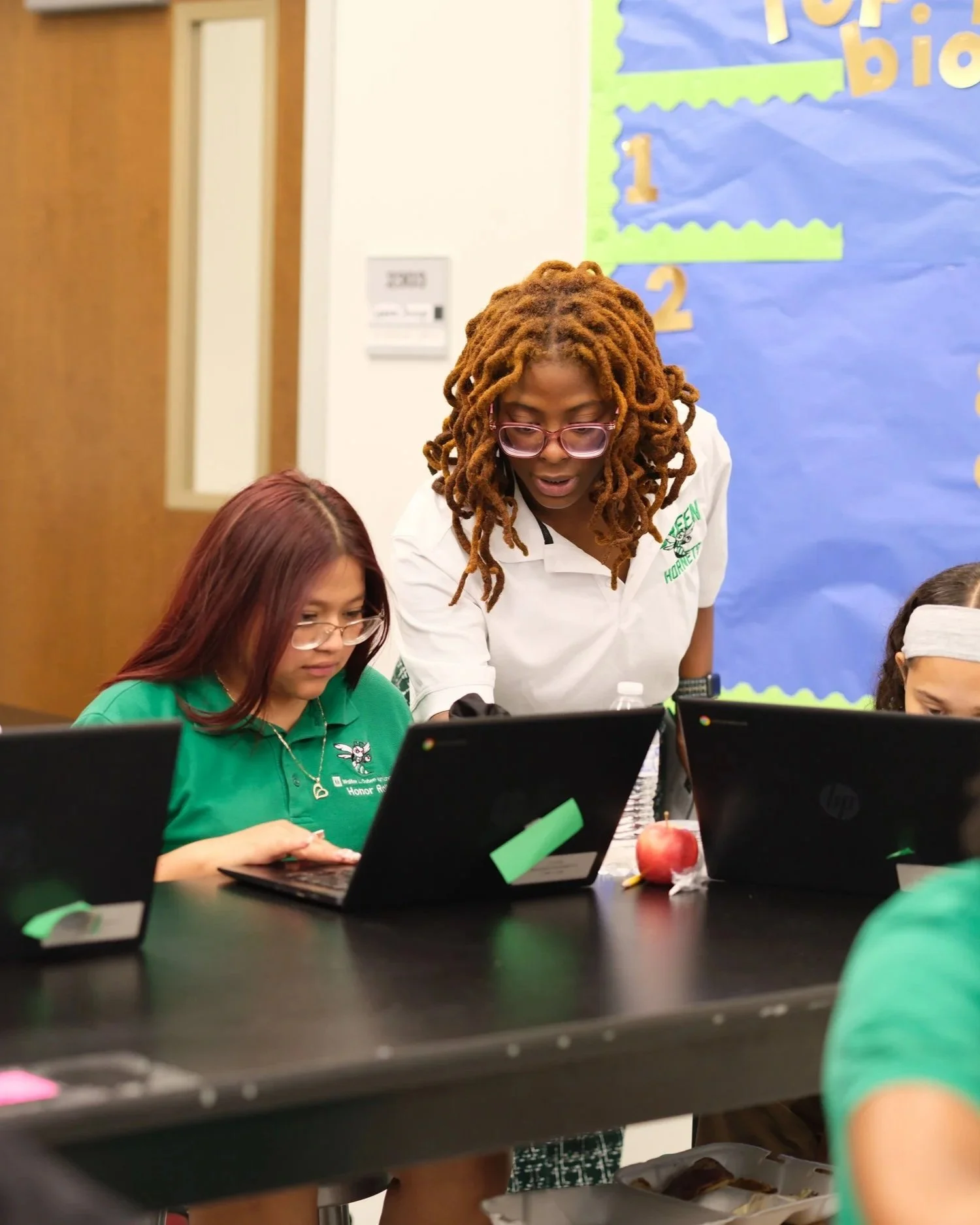 Teacher with glasses and curly hair helps students with laptops during classroom activity. High school classroom in New Orleans using AI tools. Photo courtesy of Collegiate Academies.