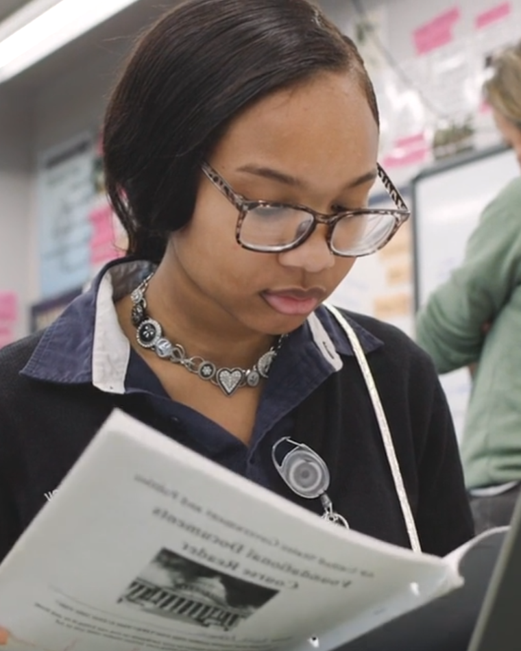 Young woman with glasses and a heart necklace reading a paper in a classroom or office setting.