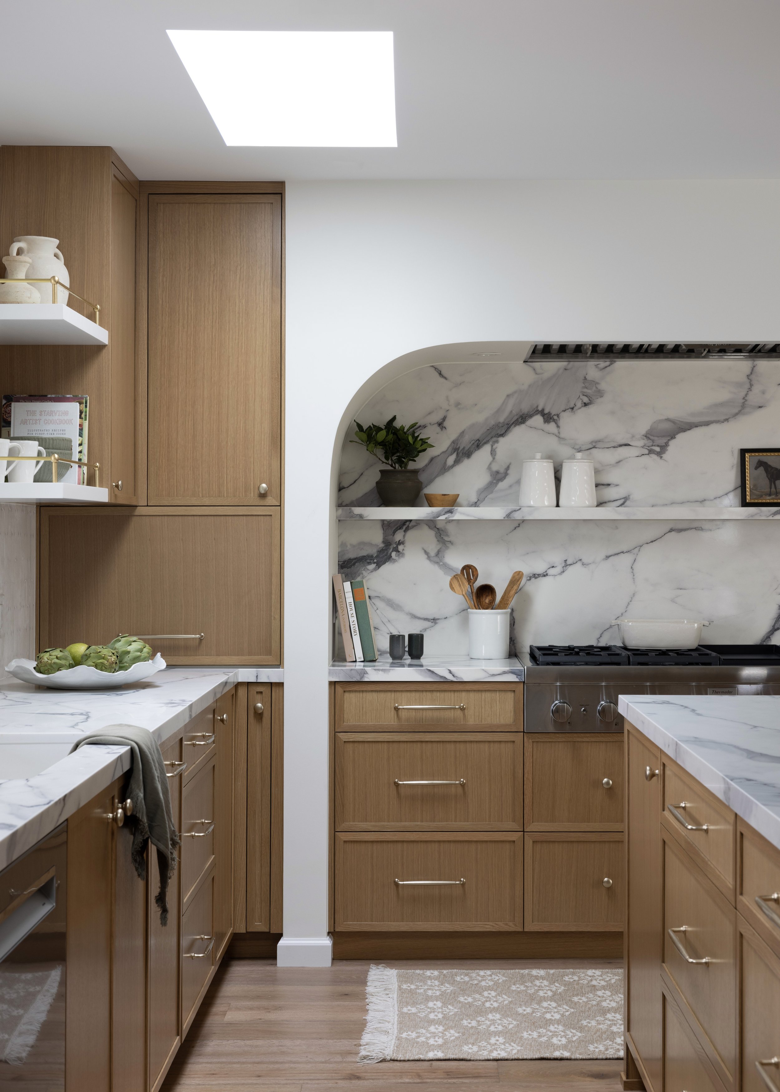 Kitchen with wooden cabinetry, marble countertops, and a marble backsplash; open shelves with dishes, a potted plant, and kitchen utensils; skylight in ceiling, beige woven rug on wooden floor.
