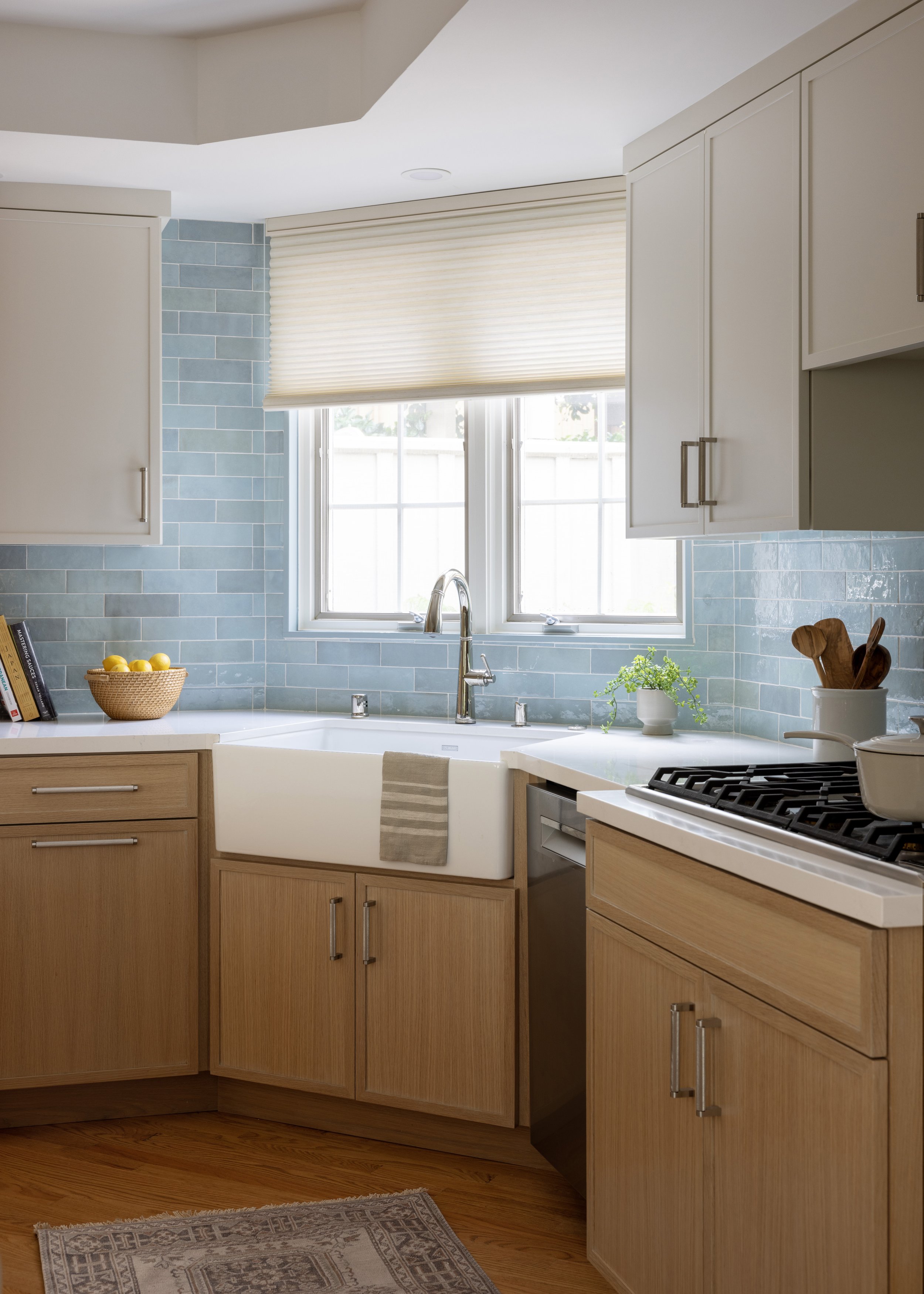 Kitchen with wooden cabinets, a white farmhouse sink, a window with a beige shade, blue subway tile backsplash, and a gas stove. There are cooking utensils and a small potted plant on the counter.