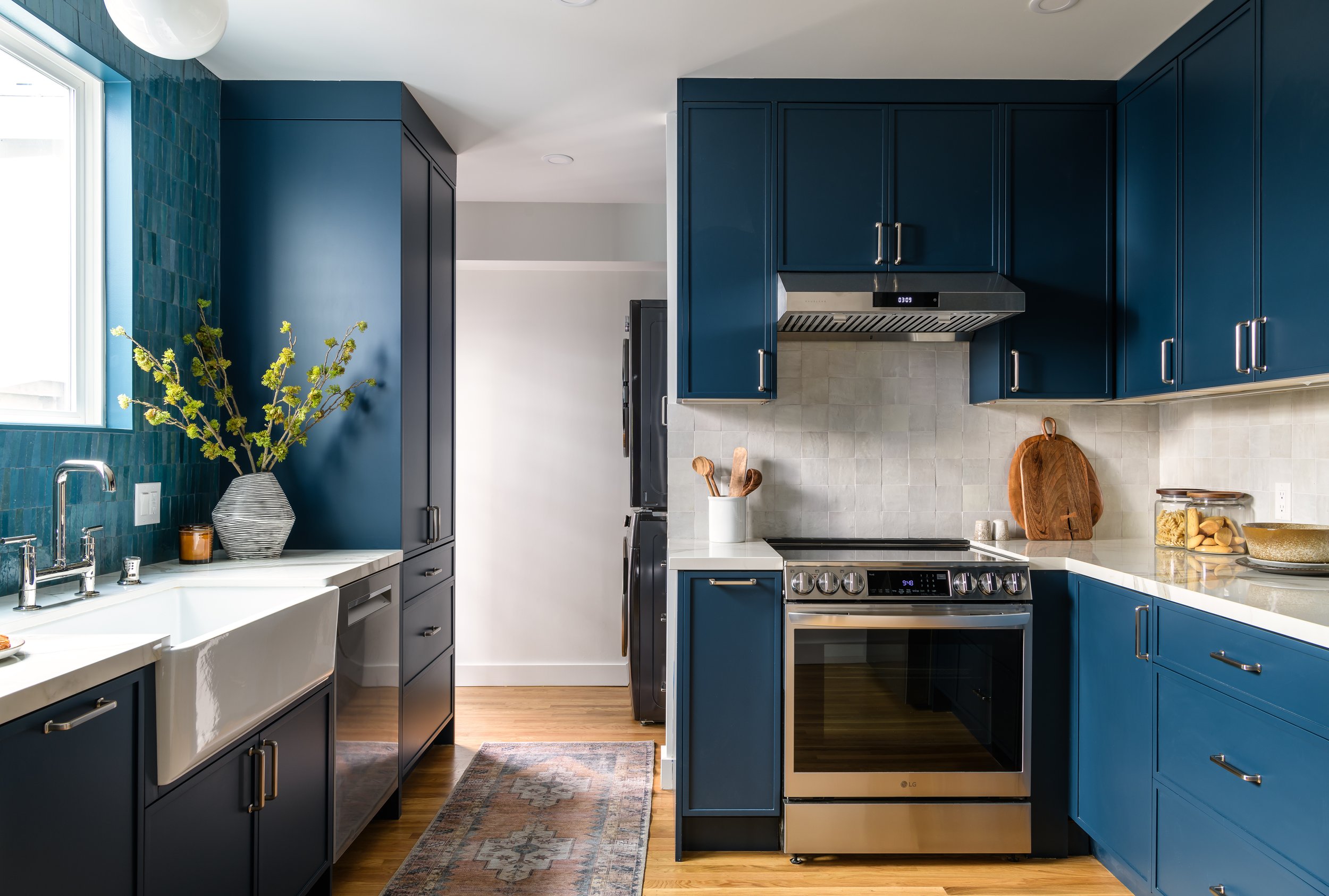 Modern kitchen with dark blue cabinets, white countertops, stainless steel oven and range hood, and a window with a view outside. Decor includes a vase with green branch, cutting boards, and jars on the counter.