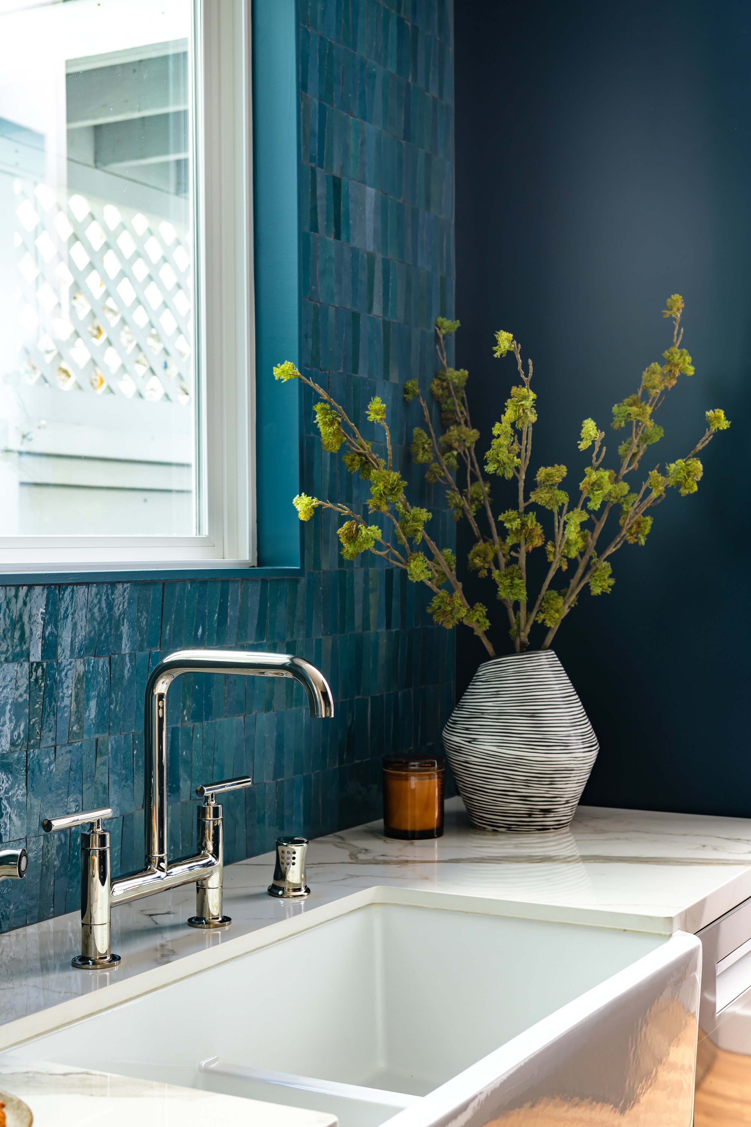 Interior view of a modern kitchen featuring a white sink with a chrome faucet, a decorative vase with a green plant, and a small jar on granite countertop. Blue tiled wall and a window with white frame in the background.