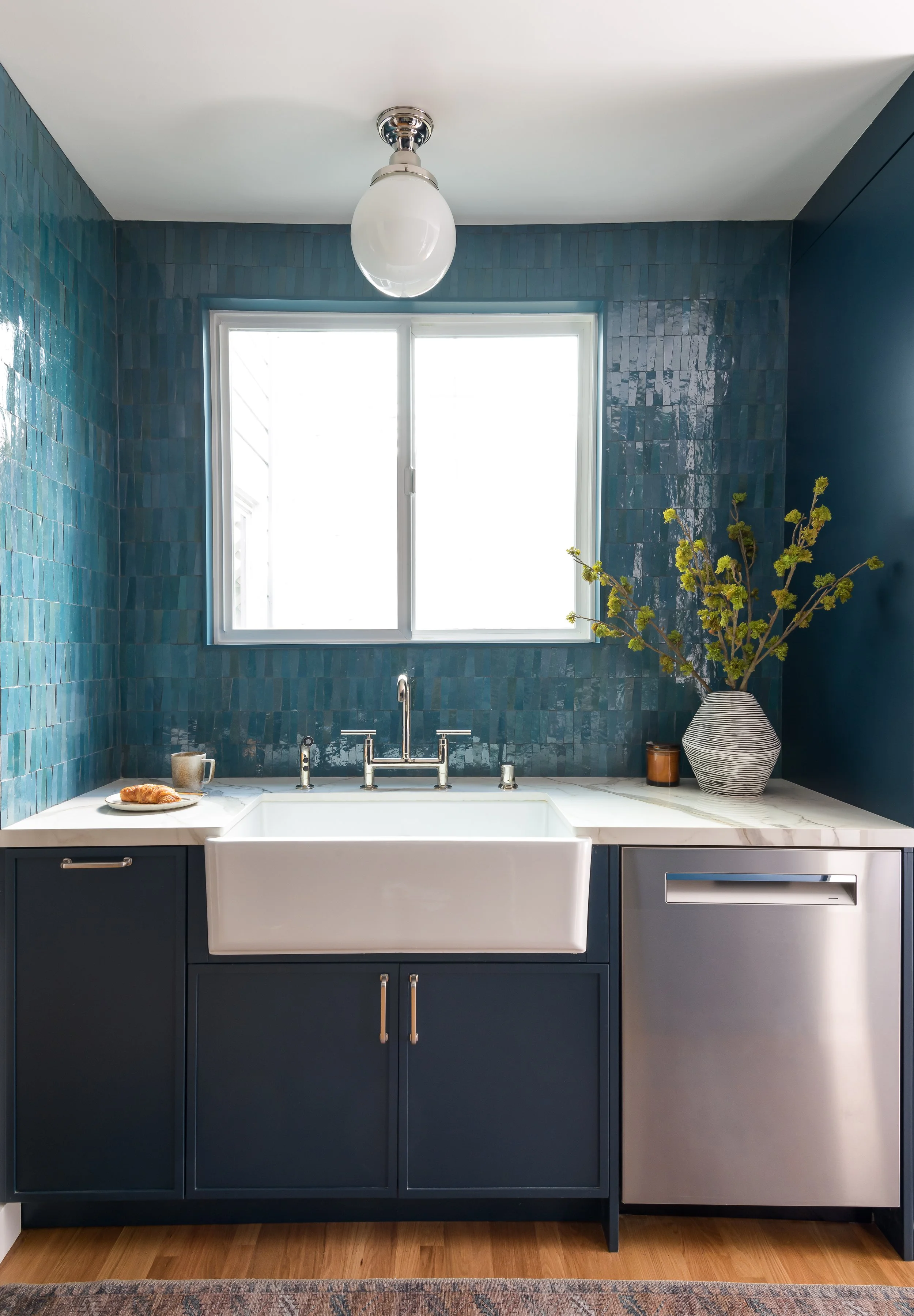 Modern kitchen with blue tiled walls, white farmhouse sink, and blue cabinetry. A window above the sink lets in natural light. On the counter, there is a croissant with a mug, a decorative vase with green branches, and a small container.