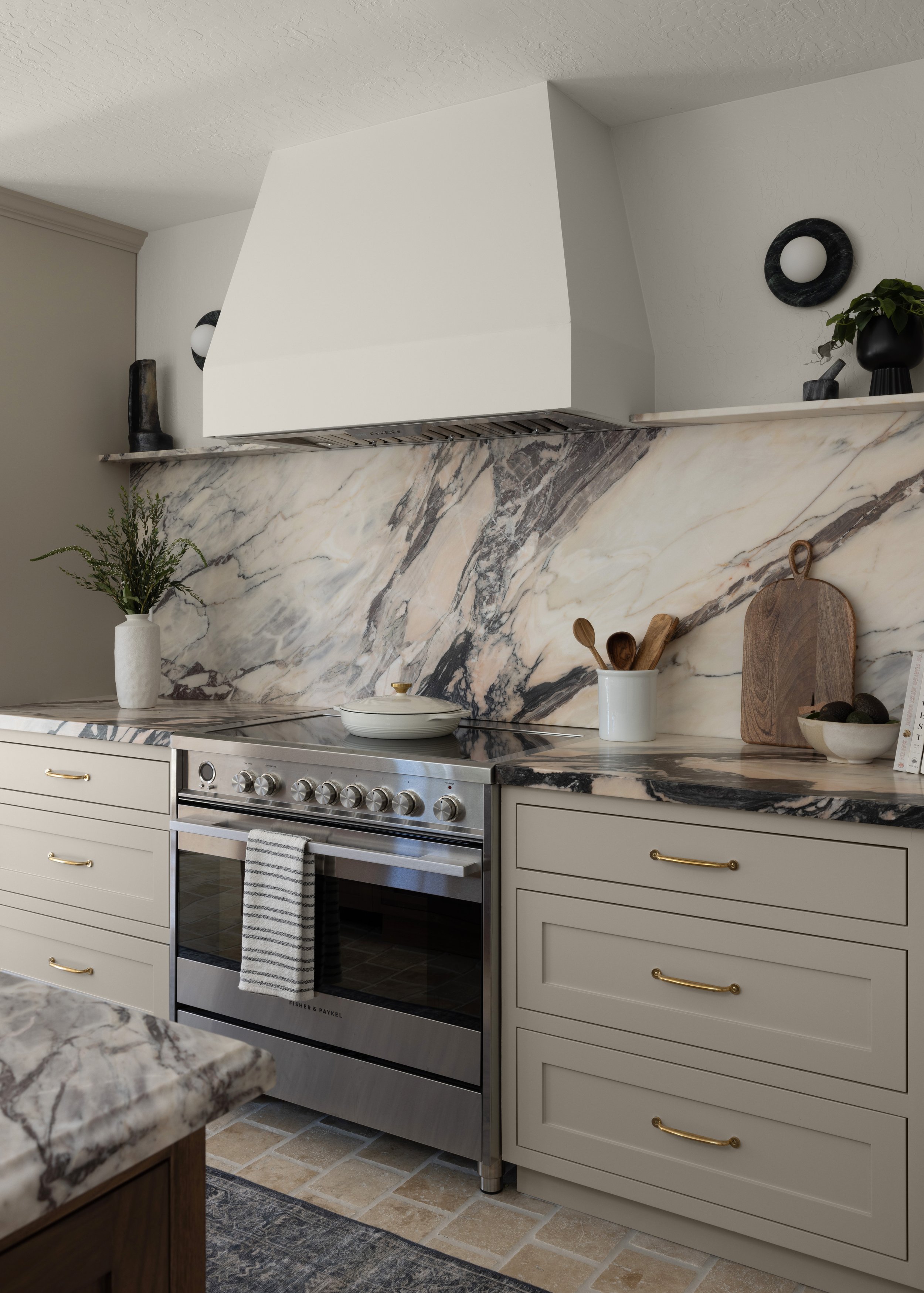 Kitchen with marble backsplash, beige cabinets with gold handles, stainless steel oven, and decorative black vases and plants.