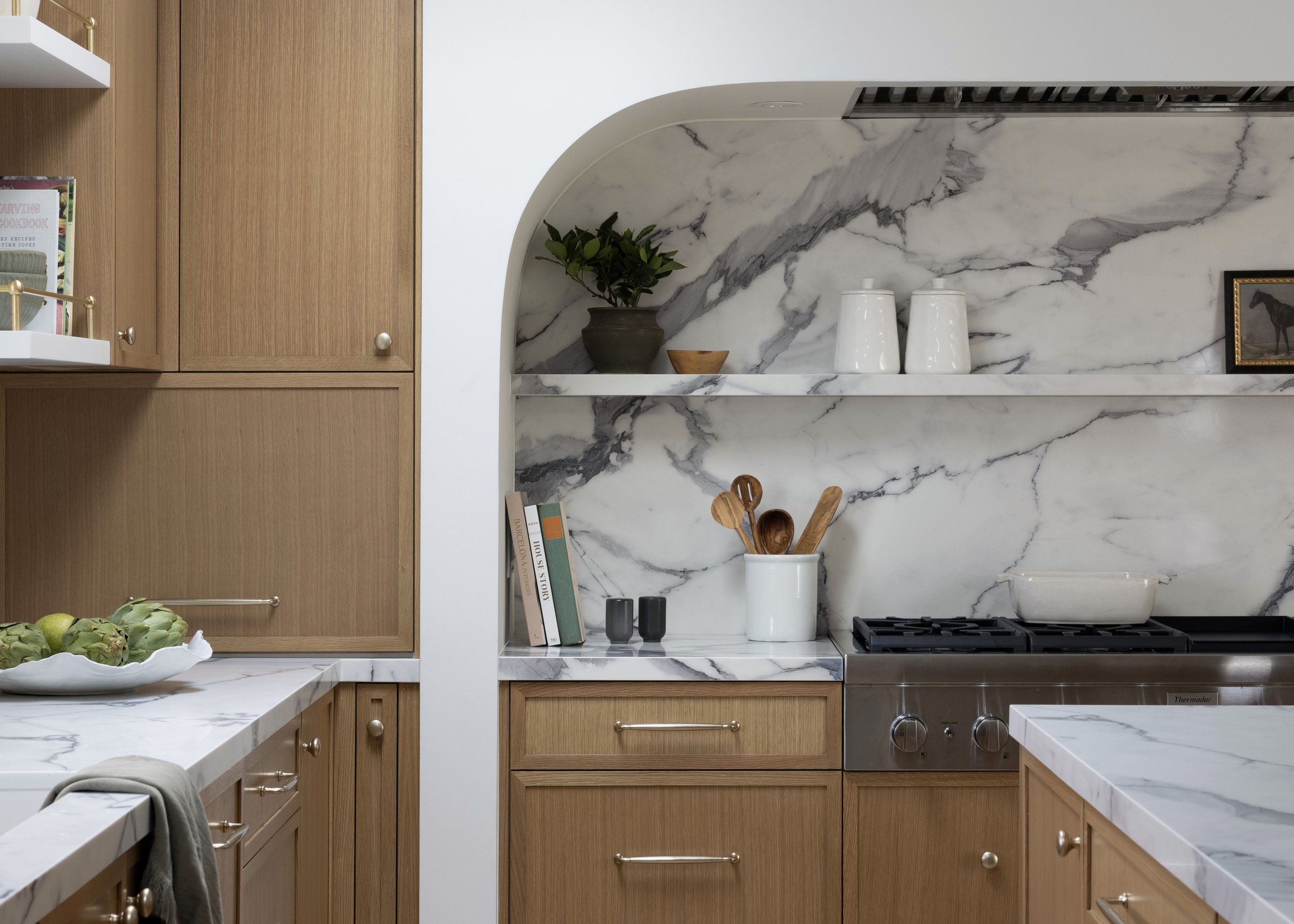Kitchen with wooden cabinets, marble backsplash, and marble countertops. There is a white ceramic utensil holder with wooden utensils, a small shelf with two white vases, a potted plant, and a loaf of bread on the counter.
