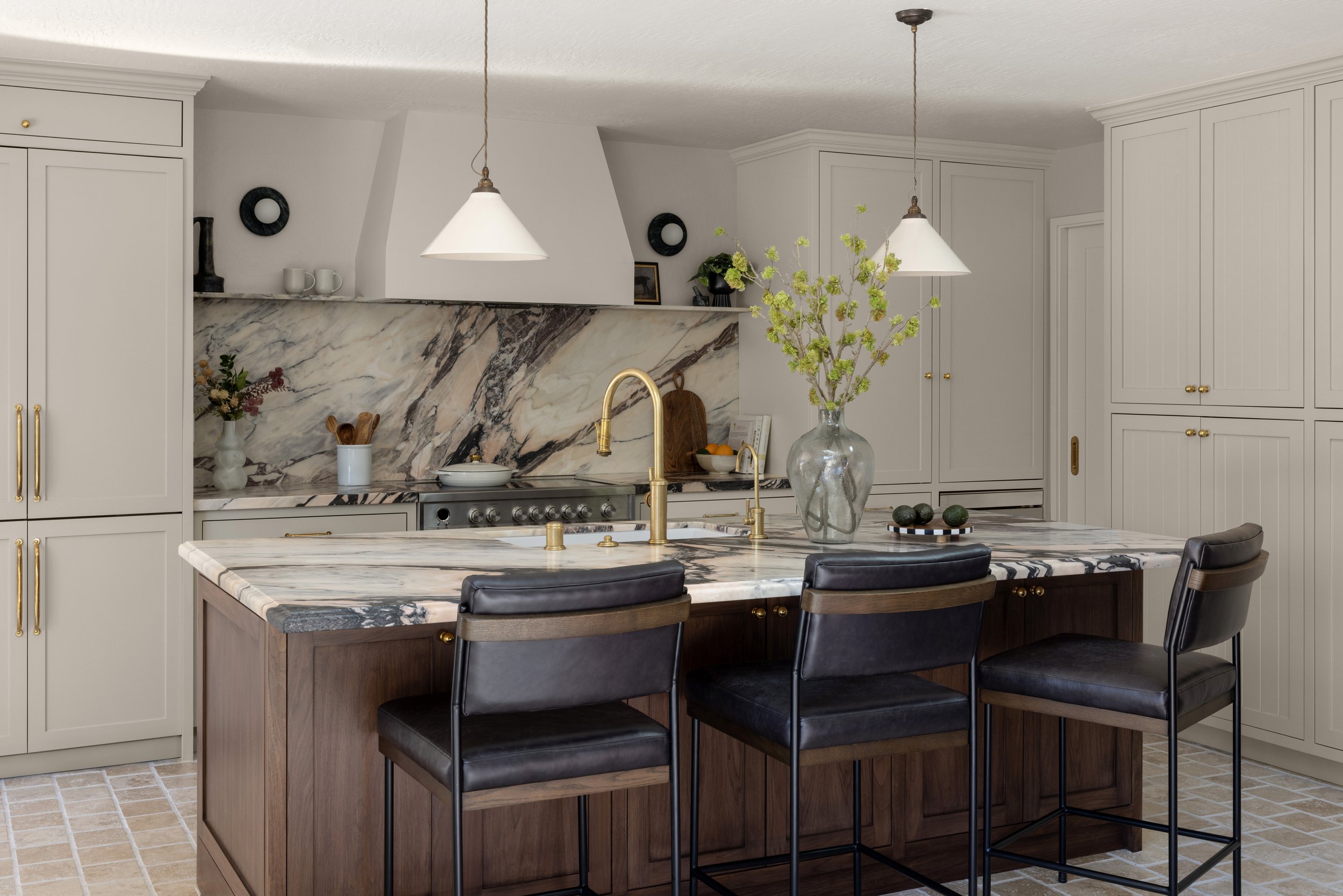 A modern kitchen with a marble island and backsplash, beige cabinets with gold handles, black barstools with leather seats, a large glass vase with green branches on the island, and pendant lights hanging from the ceiling.