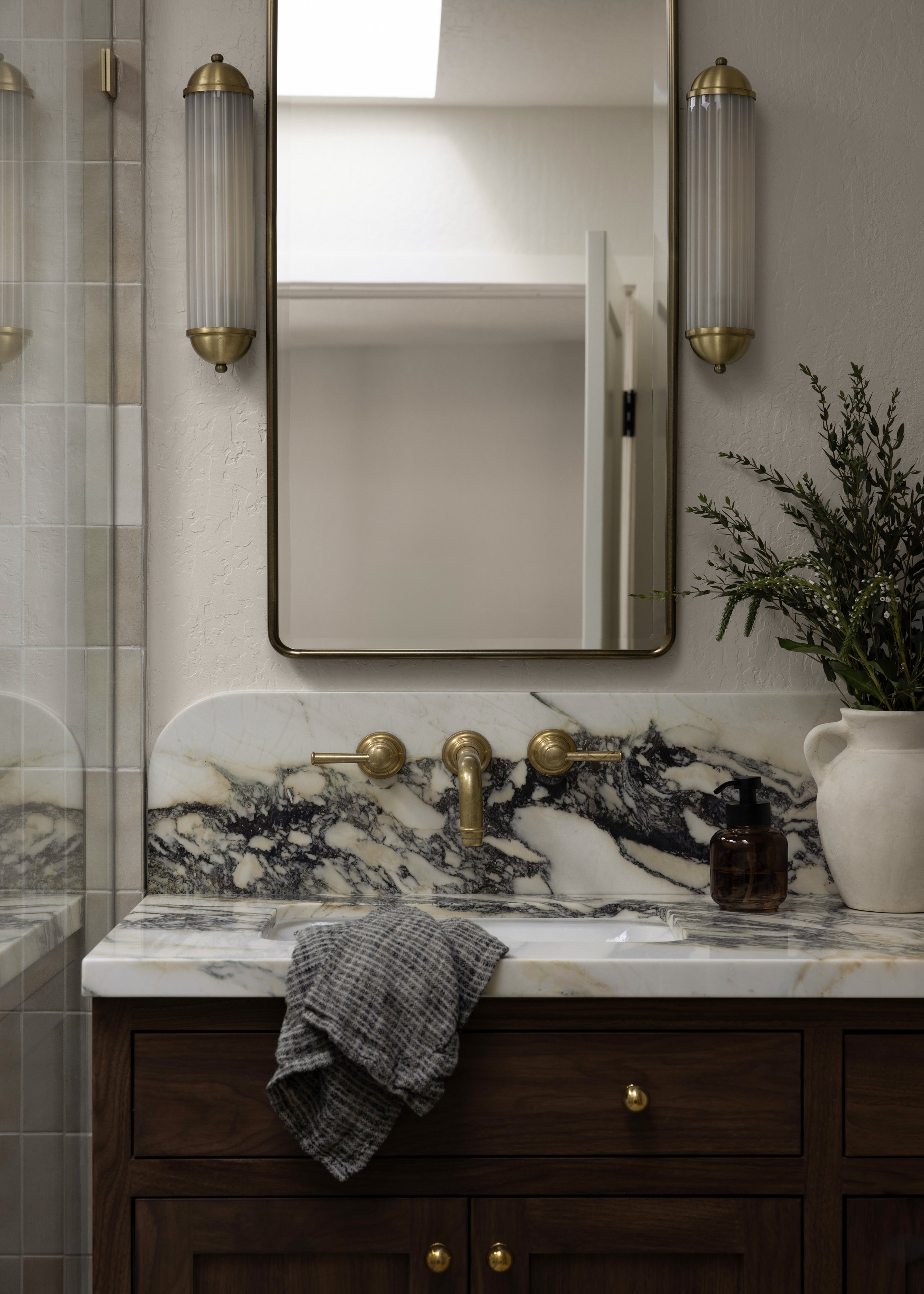 Bathroom vanity with a marble countertop, dark wood cabinet, and brass fixtures. A mirror hangs above and two wall sconces are on either side. A black soap dispenser and potted plant are on the right. A gray towel is draped over the counter.