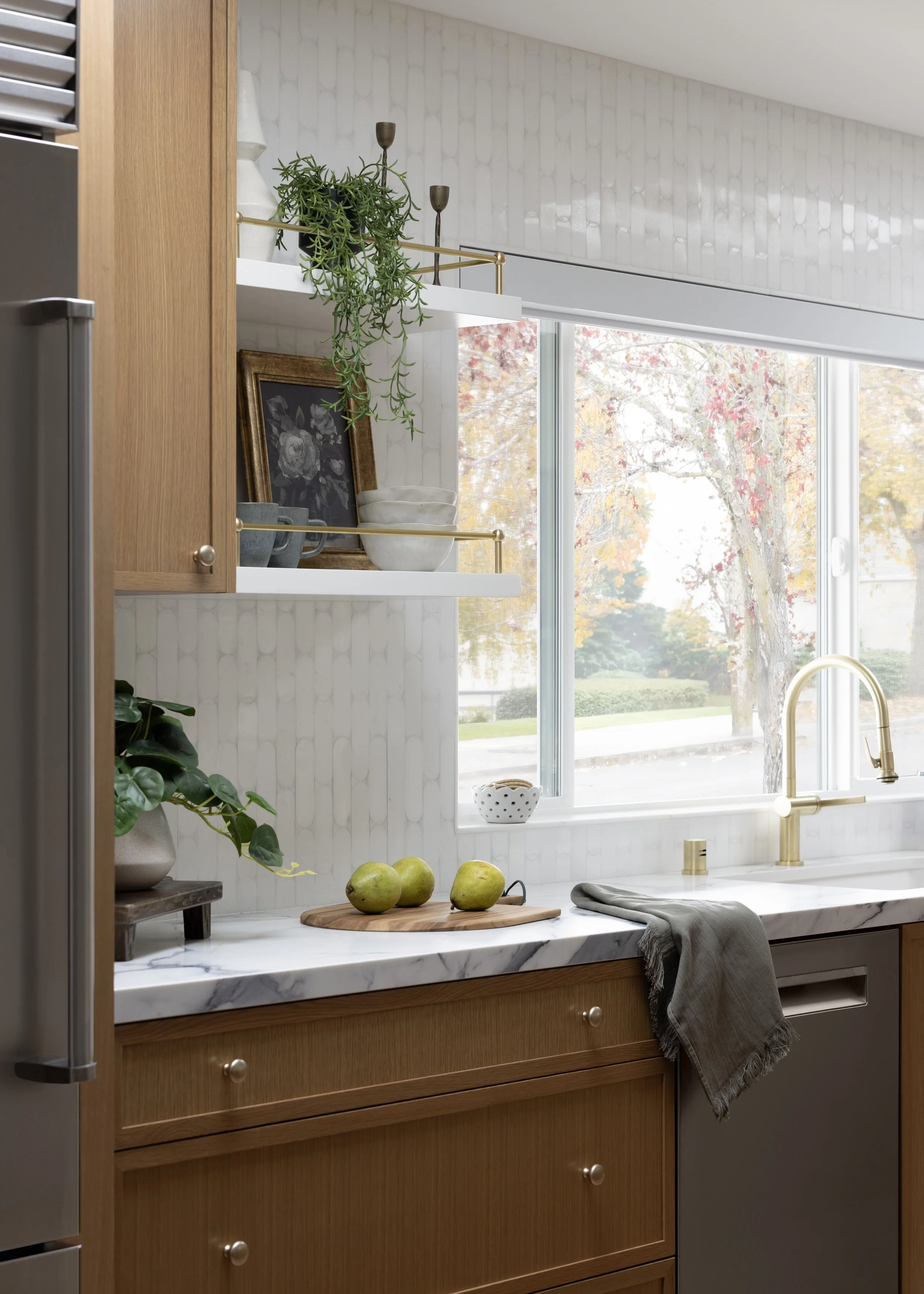 Kitchen with white marble countertop, wooden cabinets, gold faucet, and open shelves with decorative items. Green apples on a wooden cutting board, potted plant, and view outside through large window with fall foliage.
