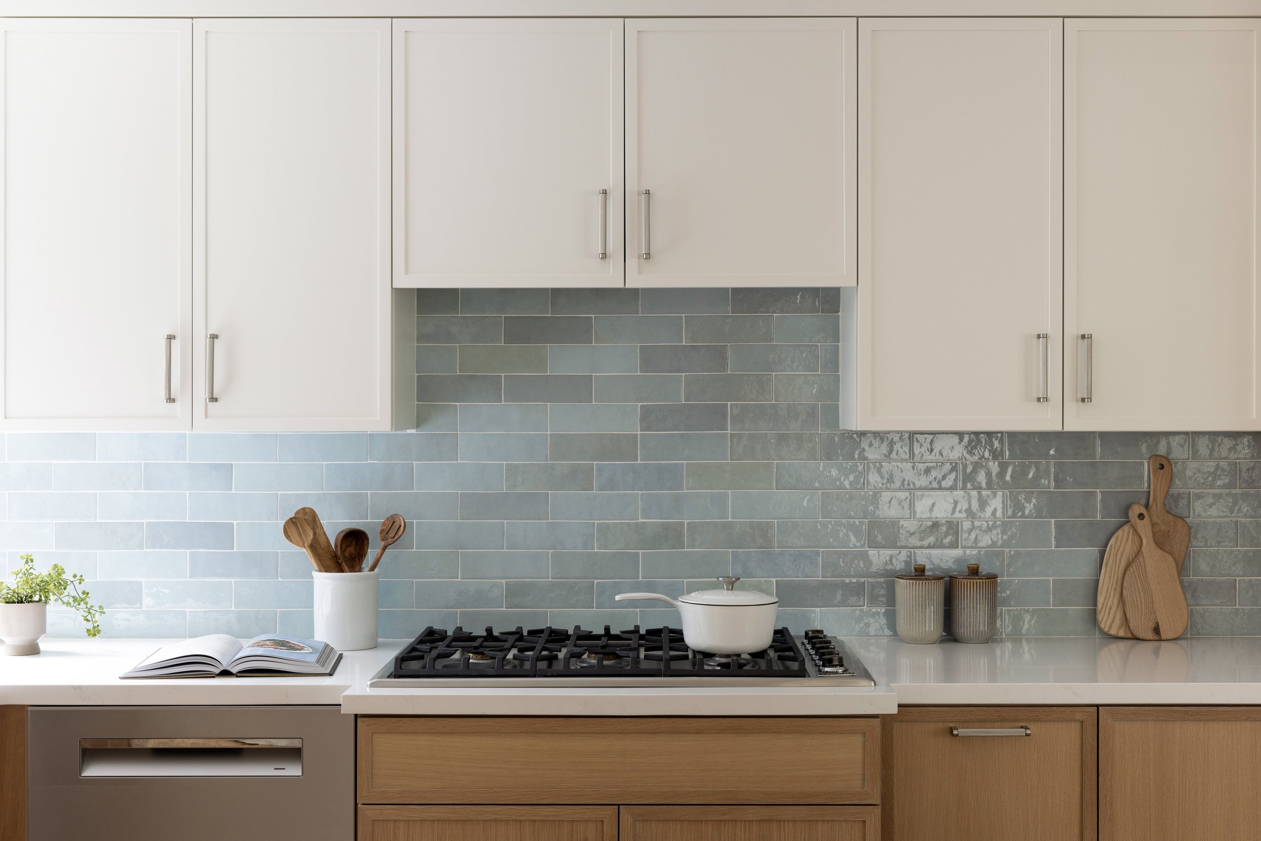 Kitchen with white cabinets, blue tile backsplash, and wooden lower cabinets. A white pot with wooden utensils, a small potted plant, an open cookbook, and two ceramic jars are on the countertop, along with a cutting board.
