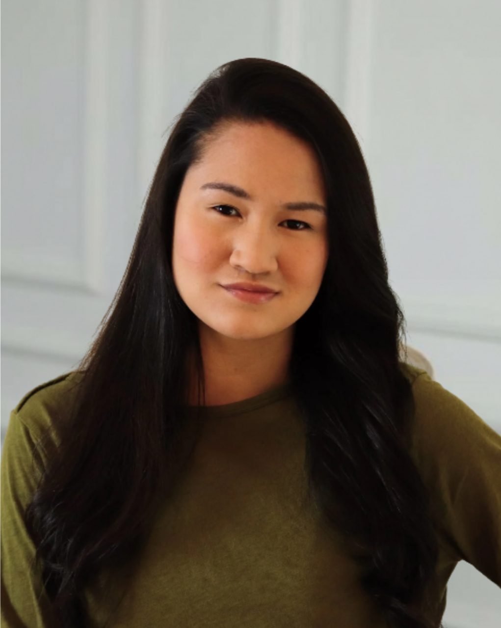 A young woman with long dark hair and light skin, wearing an olive green shirt, looking at the camera with a slight smile, standing in front of a neutral-colored wall.