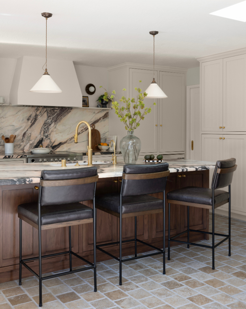 Modern kitchen with marble countertops, white cabinets, pendant lighting, black leather barstools, and a large vase with green foliage on the island.