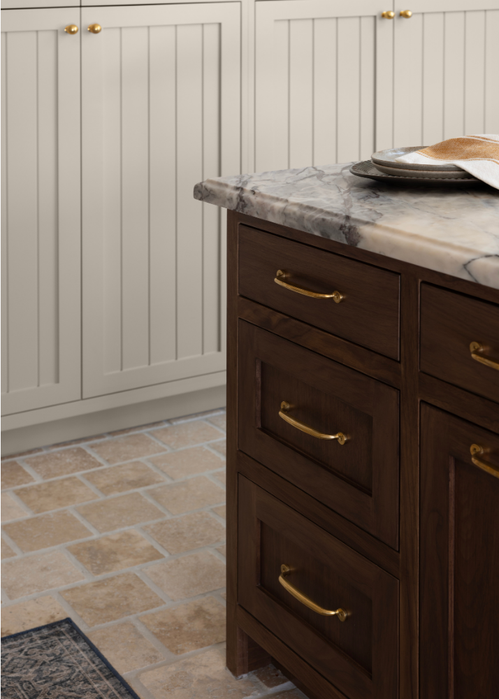 Part of a kitchen with a marble countertop and dark wooden cabinets with brass handles, beige tiled floor, and a white paneled wall with gold knobs.