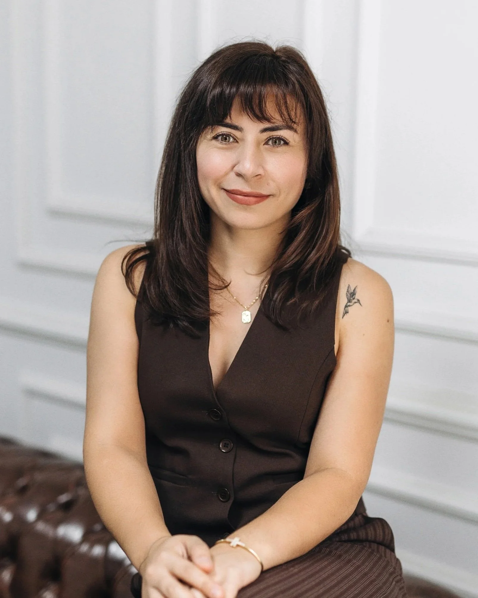 Fatma with shoulder-length dark hair, wearing a black sleeveless button-up top, sitting in front of a white paneled wall, smiling, a gold necklace, a bracelet, and a soft makeup look.